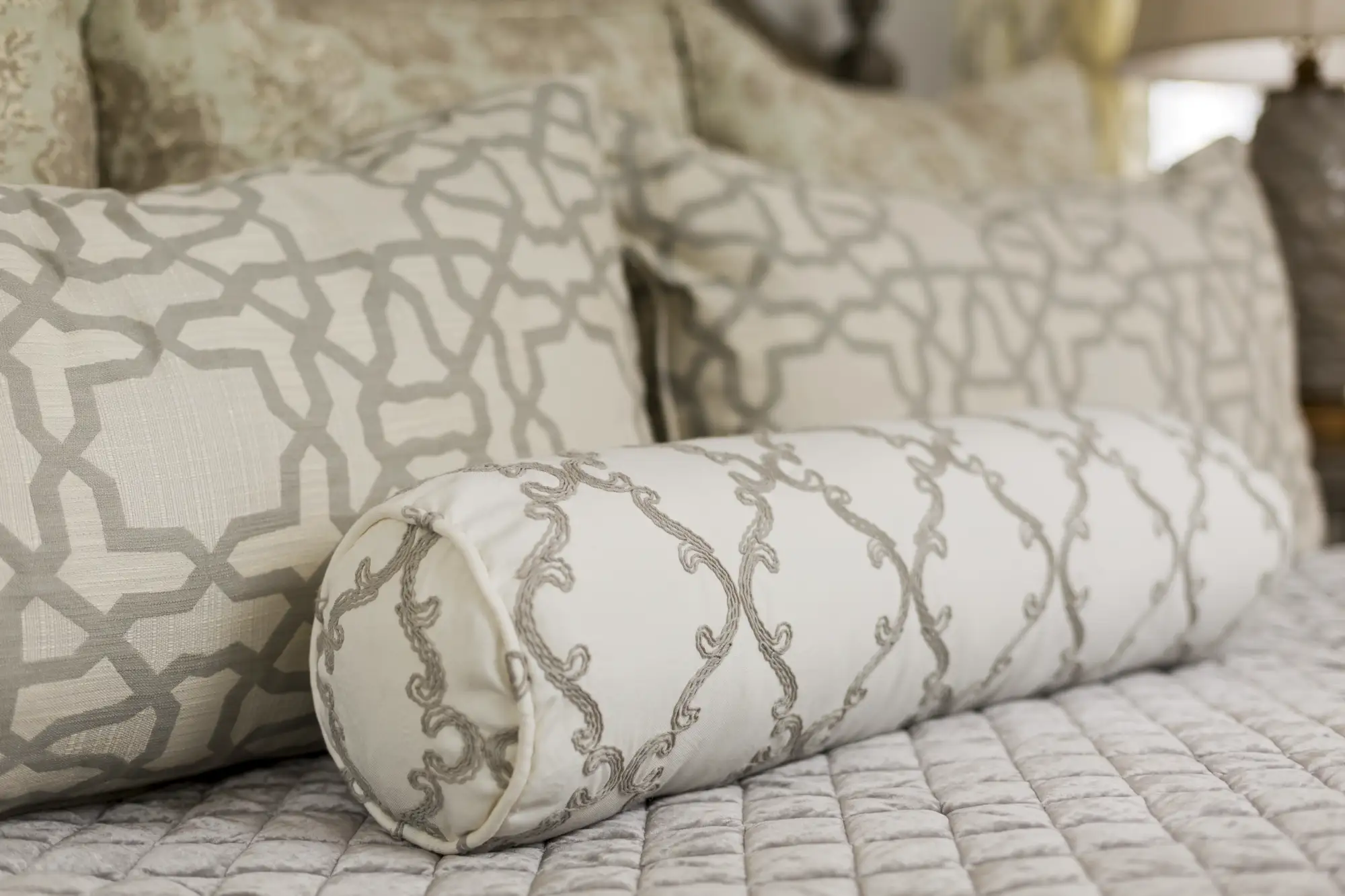 Neutral bedding detail showing a white bolster pillow embroidered with a gray scrolling pattern, set against rectangular pillows featuring a large gray geometric design on a quilted bedspread.