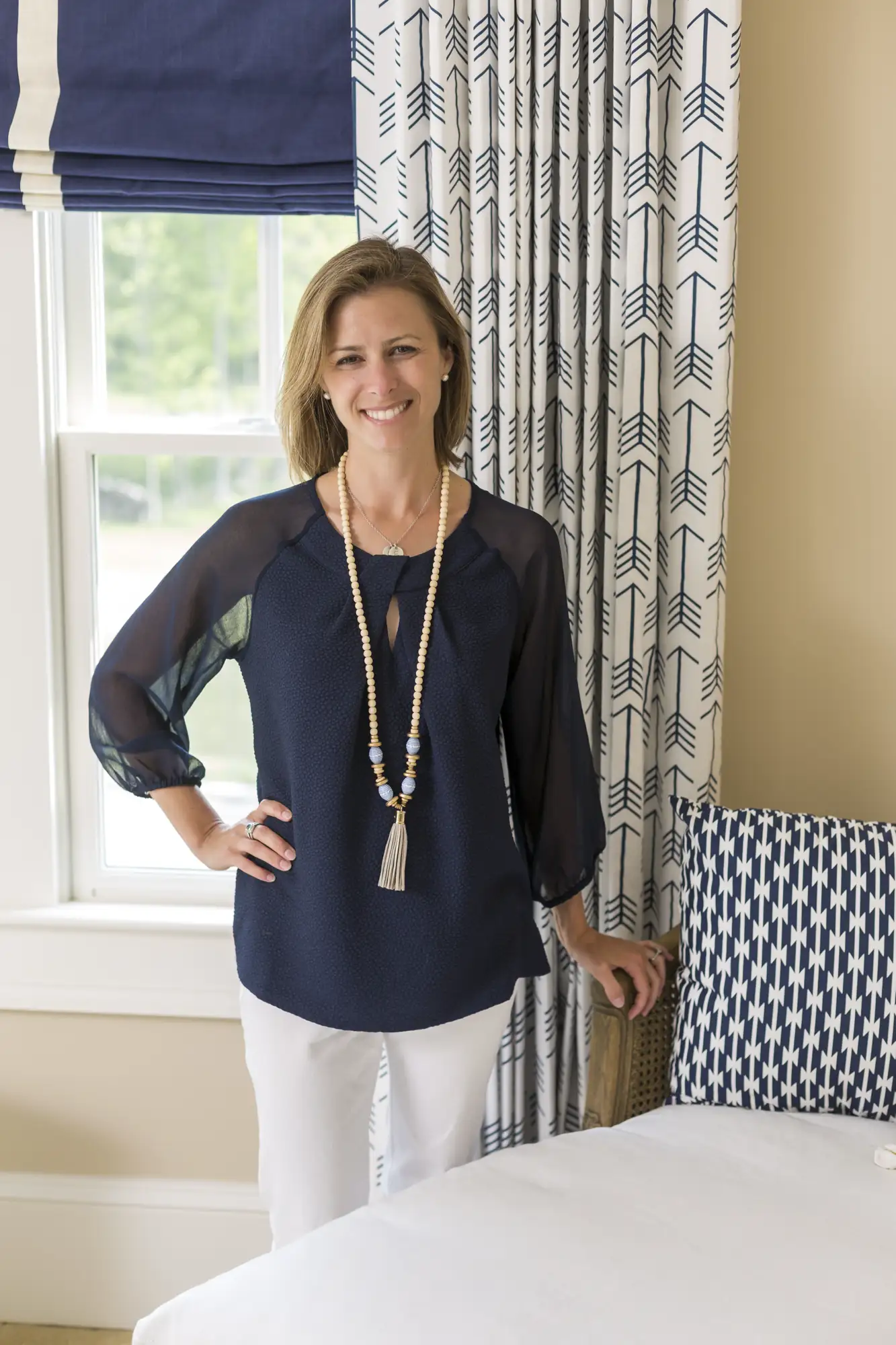 Lauren Nicole Clement smiles, wearing a navy top and beaded necklace, standing next to a window with bold blue and white patterned curtains.