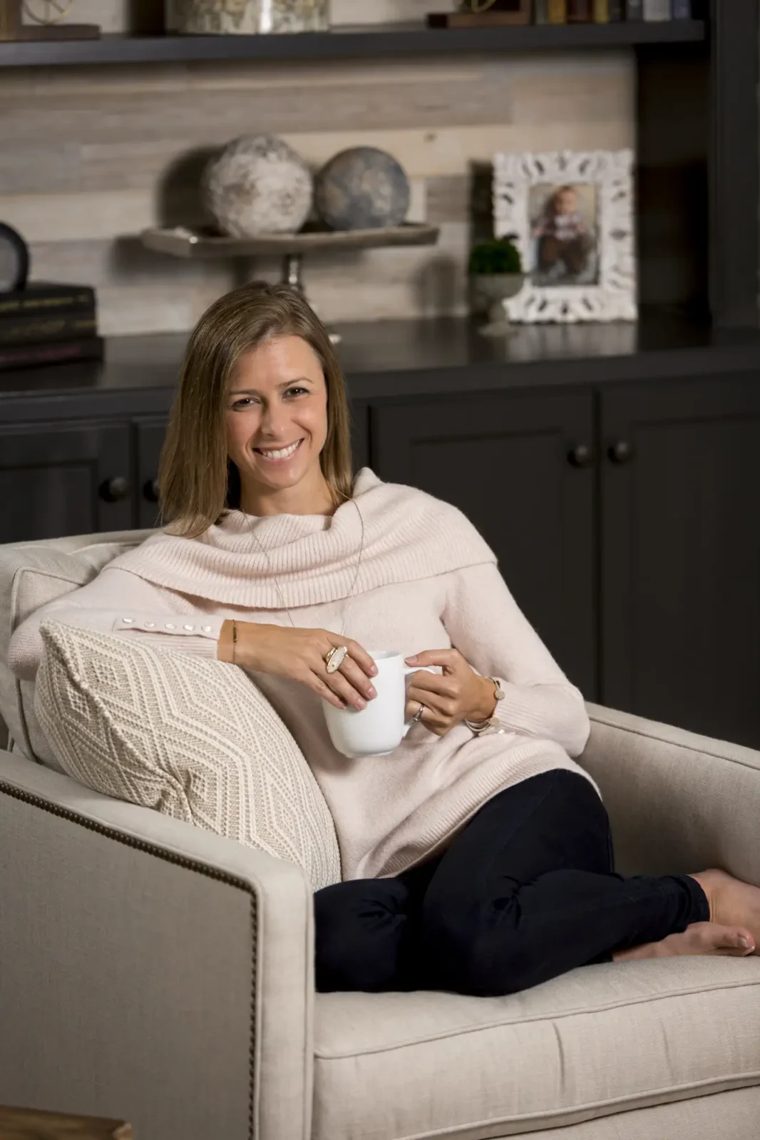 Lauren Nicole Clement sits relaxed in a beige accent chair, smiling while holding a white mug. She wears a pink cowl-neck sweater.