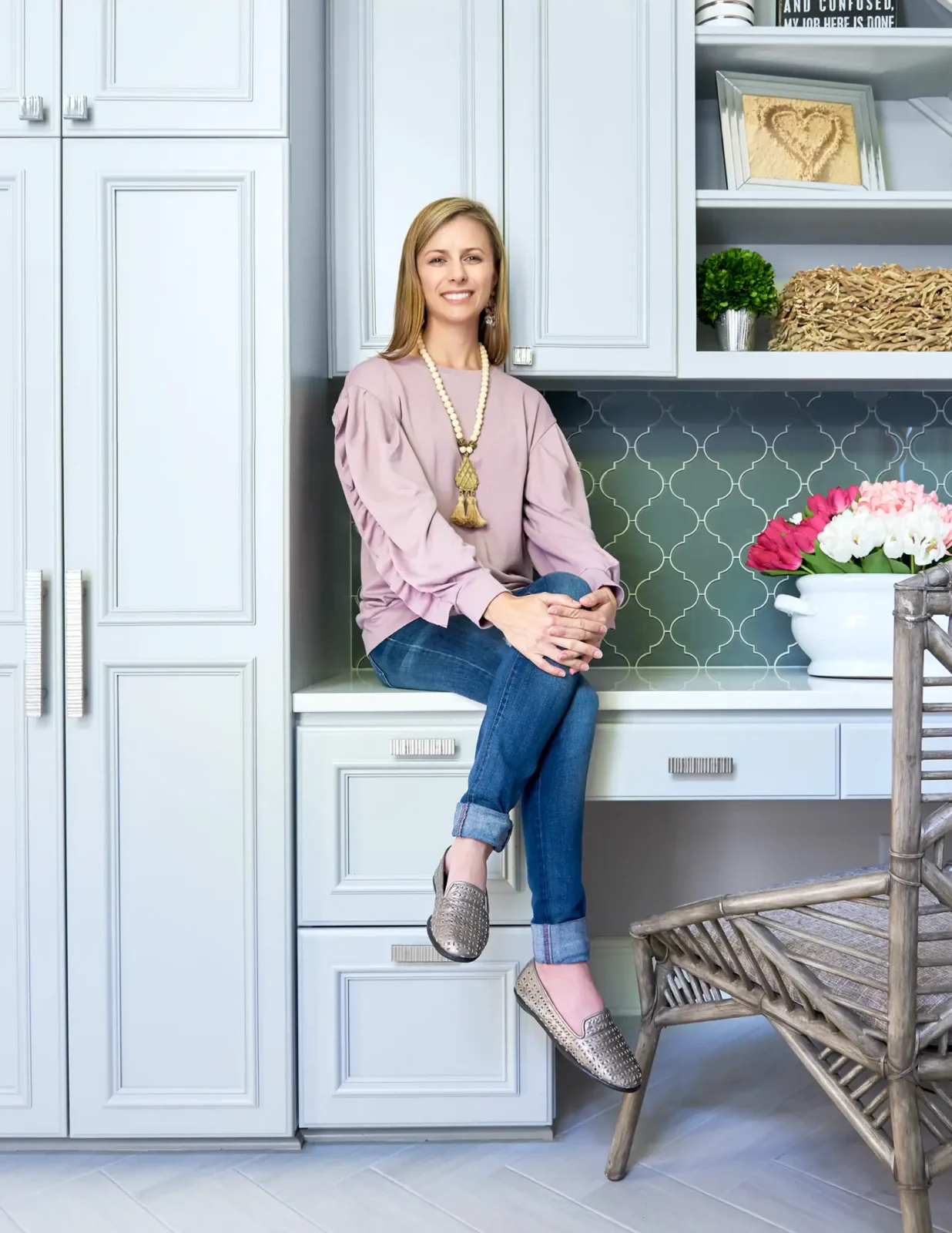 Lauren Nicole Clement, wearing a mauve top and jeans, sits smiling on a built-in desk counter against a decorative gray tile backsplash and light cabinets.