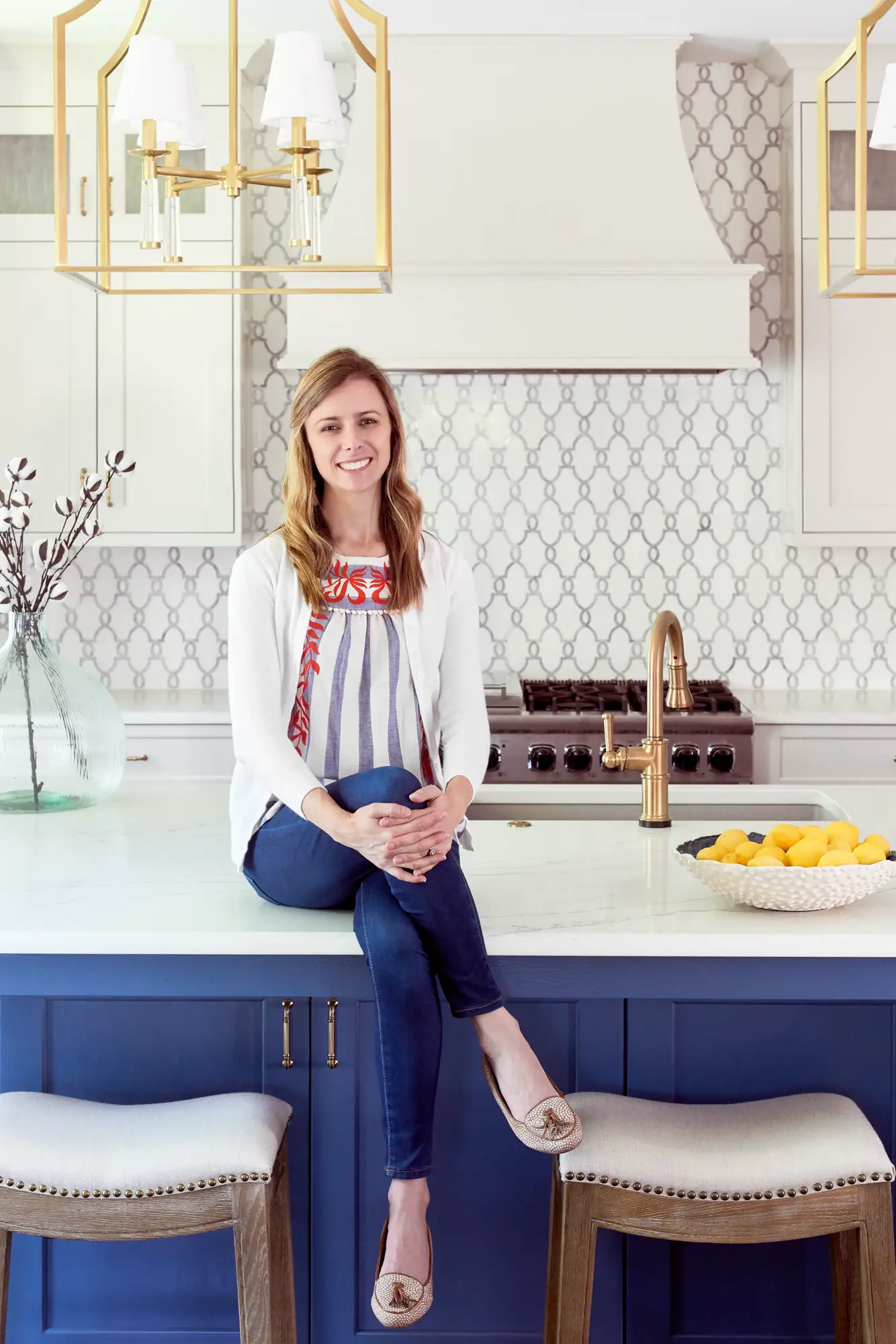 Interior designer Lauren Nicole Clement sits on a white kitchen counter, smiling confidently. The modern kitchen features a navy island, brass fixtures, and a patterned gray backsplash.