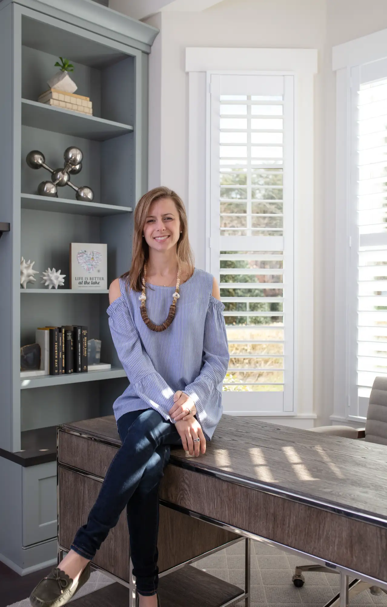 Lauren Nicole Clement sits smiling on a modern office desk. She wears a striped shirt and a wooden necklace, next to a gray bookshelf and bright, shuttered windows.