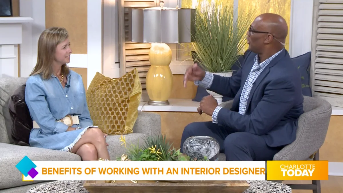 An interior designer in a light blue dress smiles while talking with a male host in a suit. He is gesturing during their conversation on a comfortable studio set.