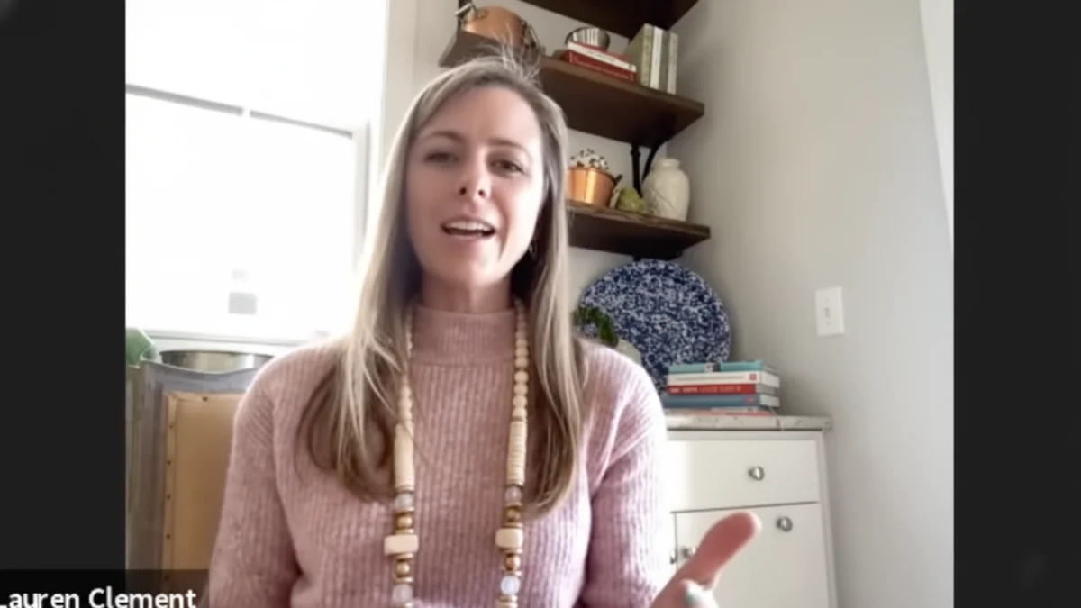 Nicole Clement in a pink sweater and beaded necklace speaks during a video call, gesturing with her hand in front of stylish floating shelves.