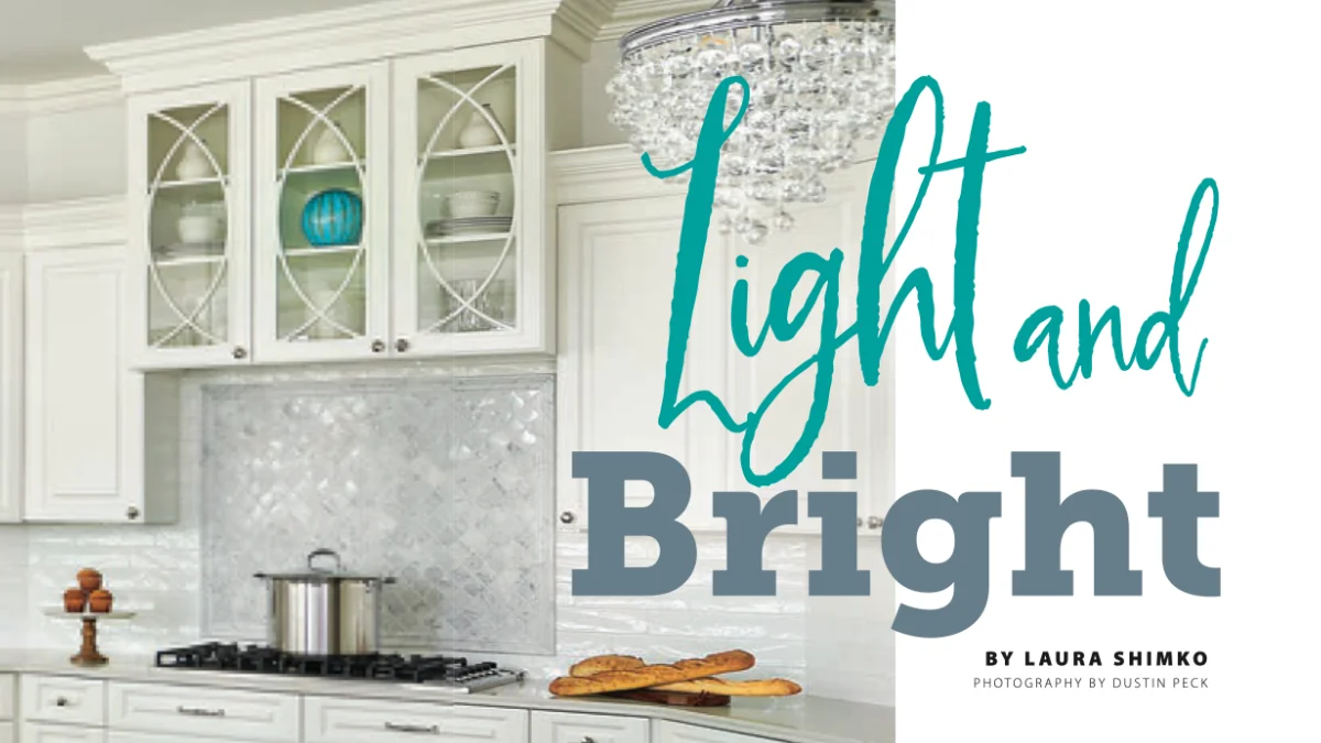 An elegant white kitchen installation with glass-front cabinets, a mosaic backsplash, a crystal chandelier, and baguettes on the marble counter.