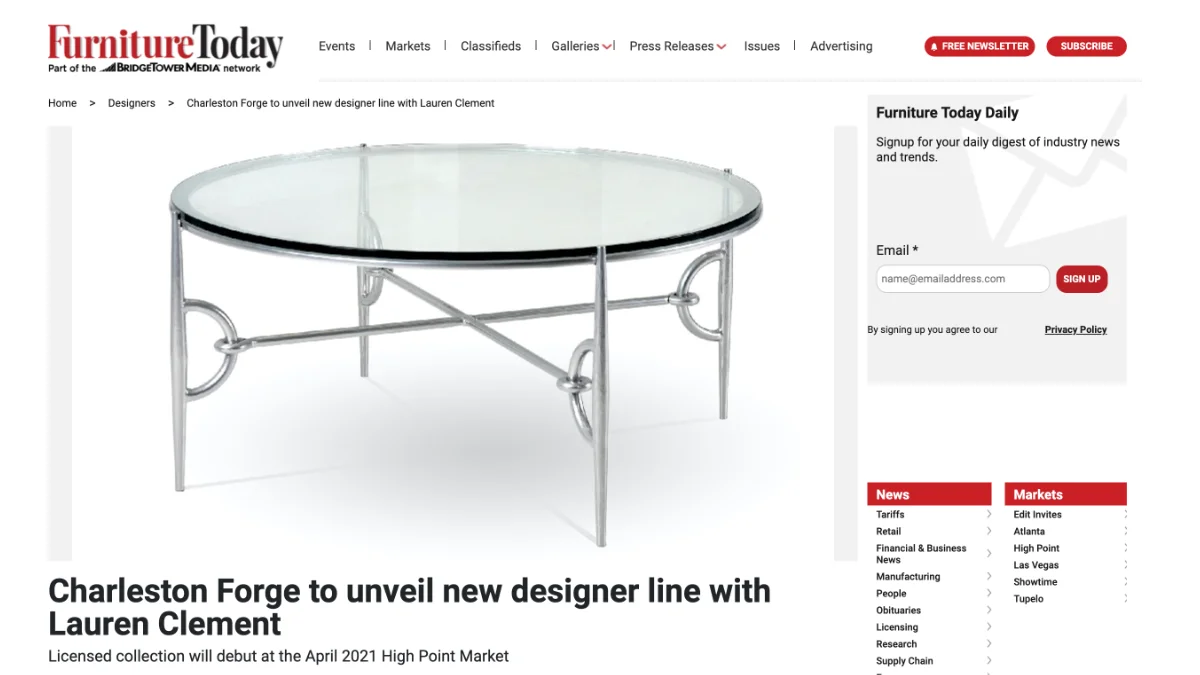 A screenshot of a website article showing a stylish round cocktail table with a clear glass top set on a polished silver metal base featuring ornate, decorative looped leg supports.