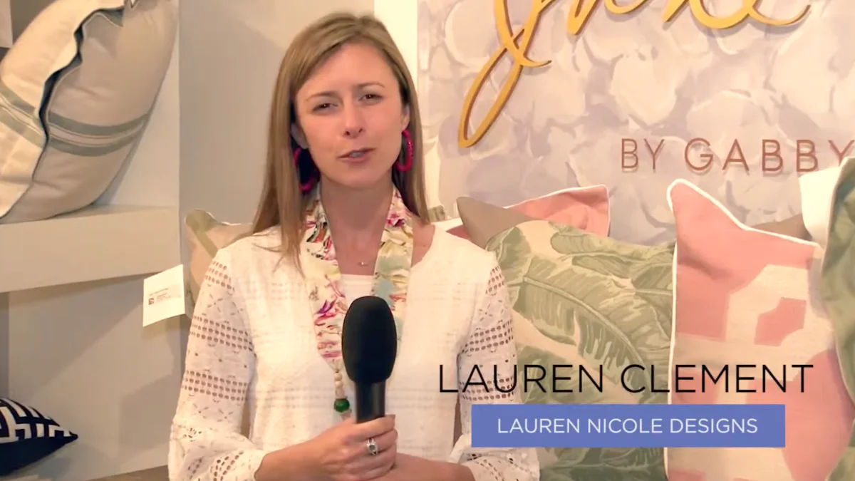 Lauren Clement, an interior designer, speaks into a microphone in a bright showroom, surrounded by patterned pillows and home decor.