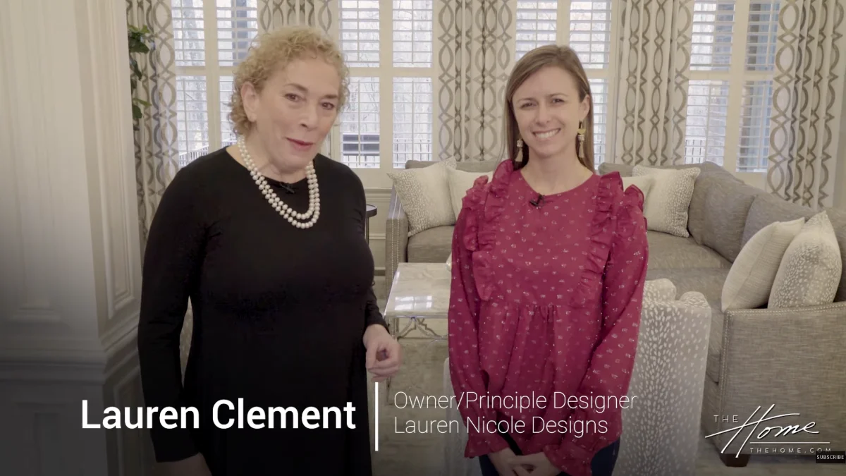 Principle Designer Lauren Nicole smiles next to The Home host in a modern living room featuring a plush gray sofa, accent chairs, and plantation shutter windows.