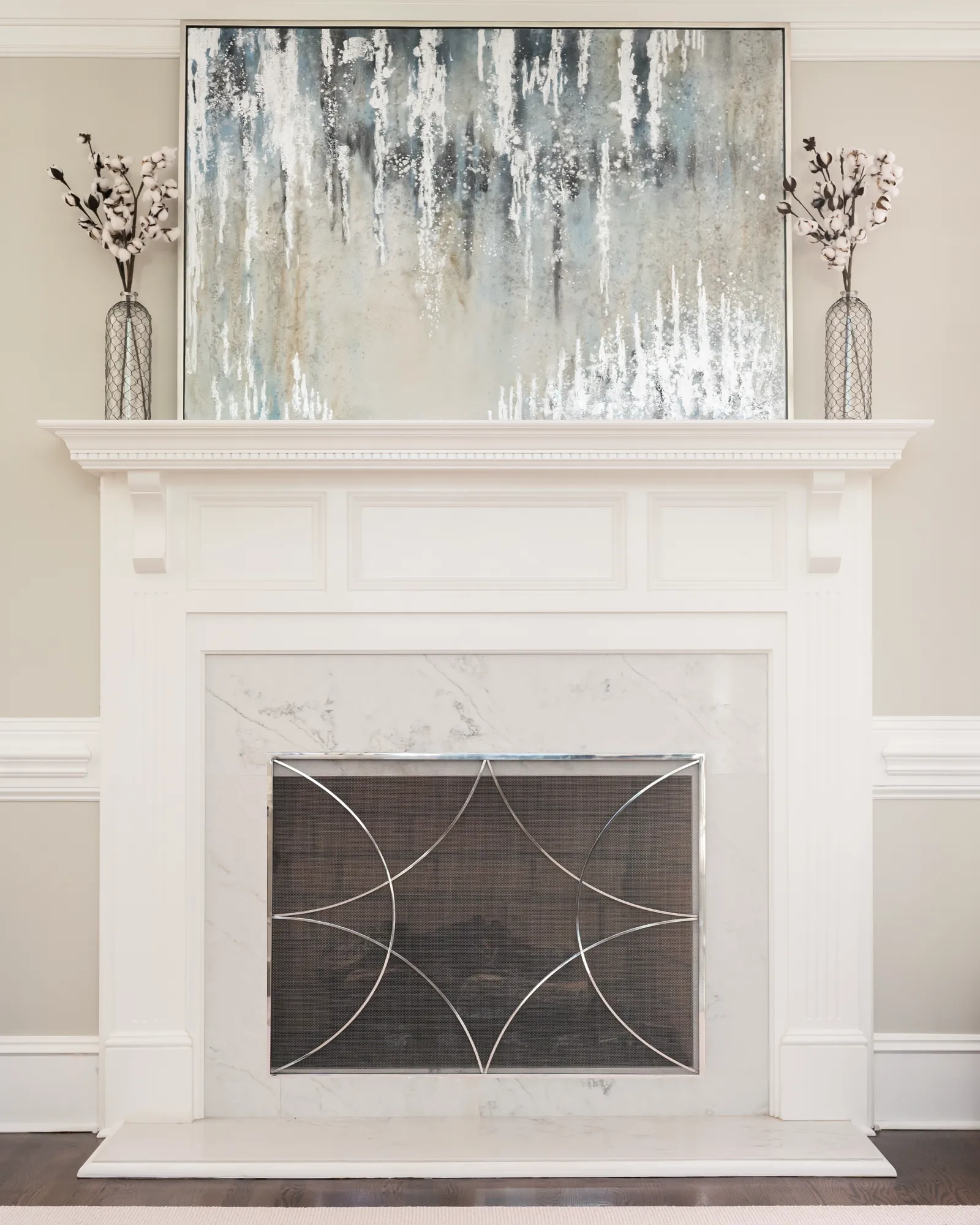 A white wooden fireplace with a marble surround and a contemporary metal screen. The mantel features a large abstract painting and two vases with cotton branches.