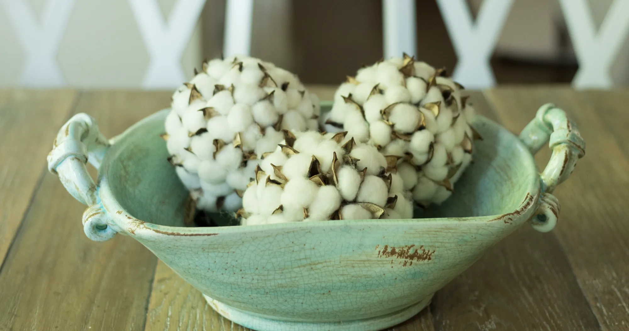 Three fluffy white decorative cotton bolls rest inside a shallow, rustic, crackled aqua ceramic bowl on a wooden table.