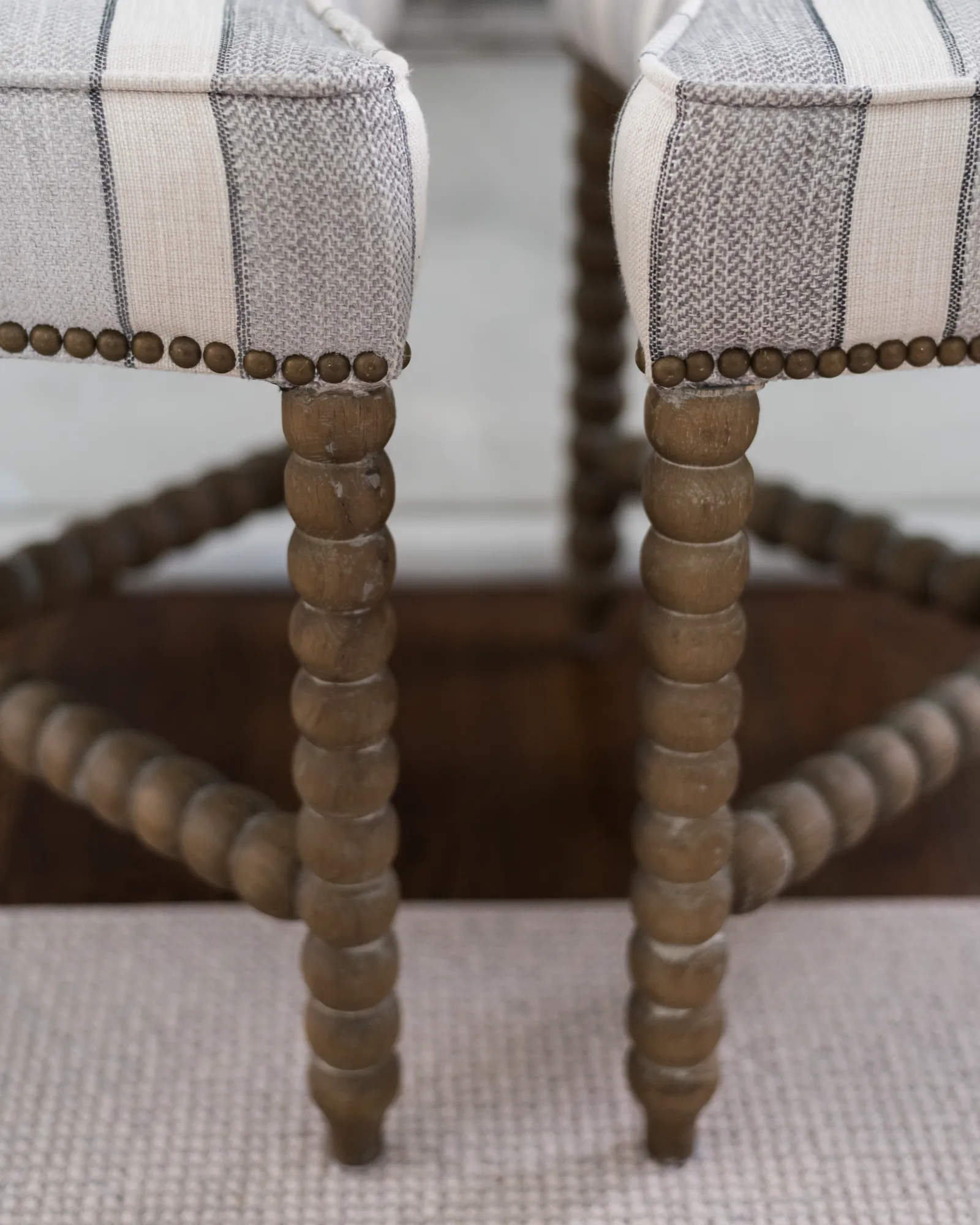 A pair of stools featuring light gray striped upholstery accented with antique nailhead trim, resting on dark, bobbin-turned wooden legs.