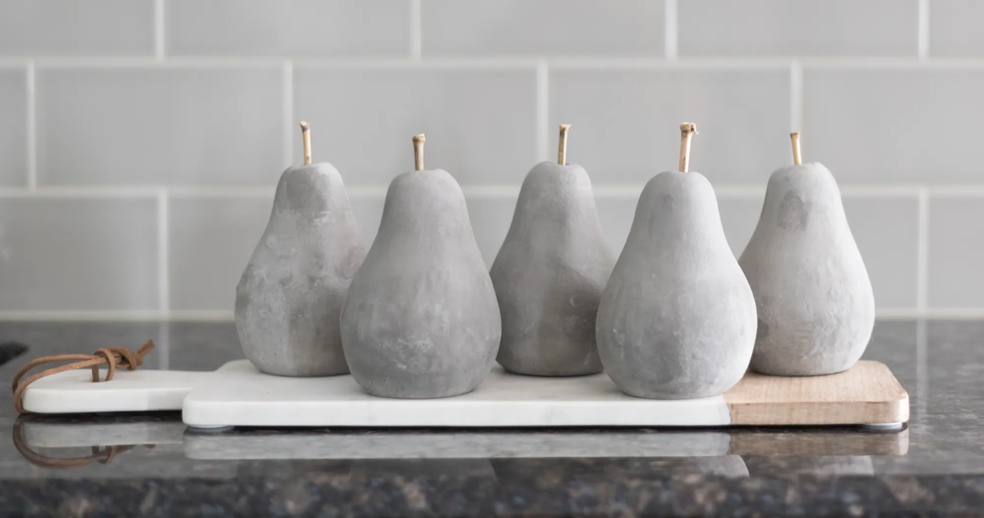 Five decorative gray stone pears with wooden stems are lined up on a marble and wood serving board against a light gray tiled kitchen backsplash.