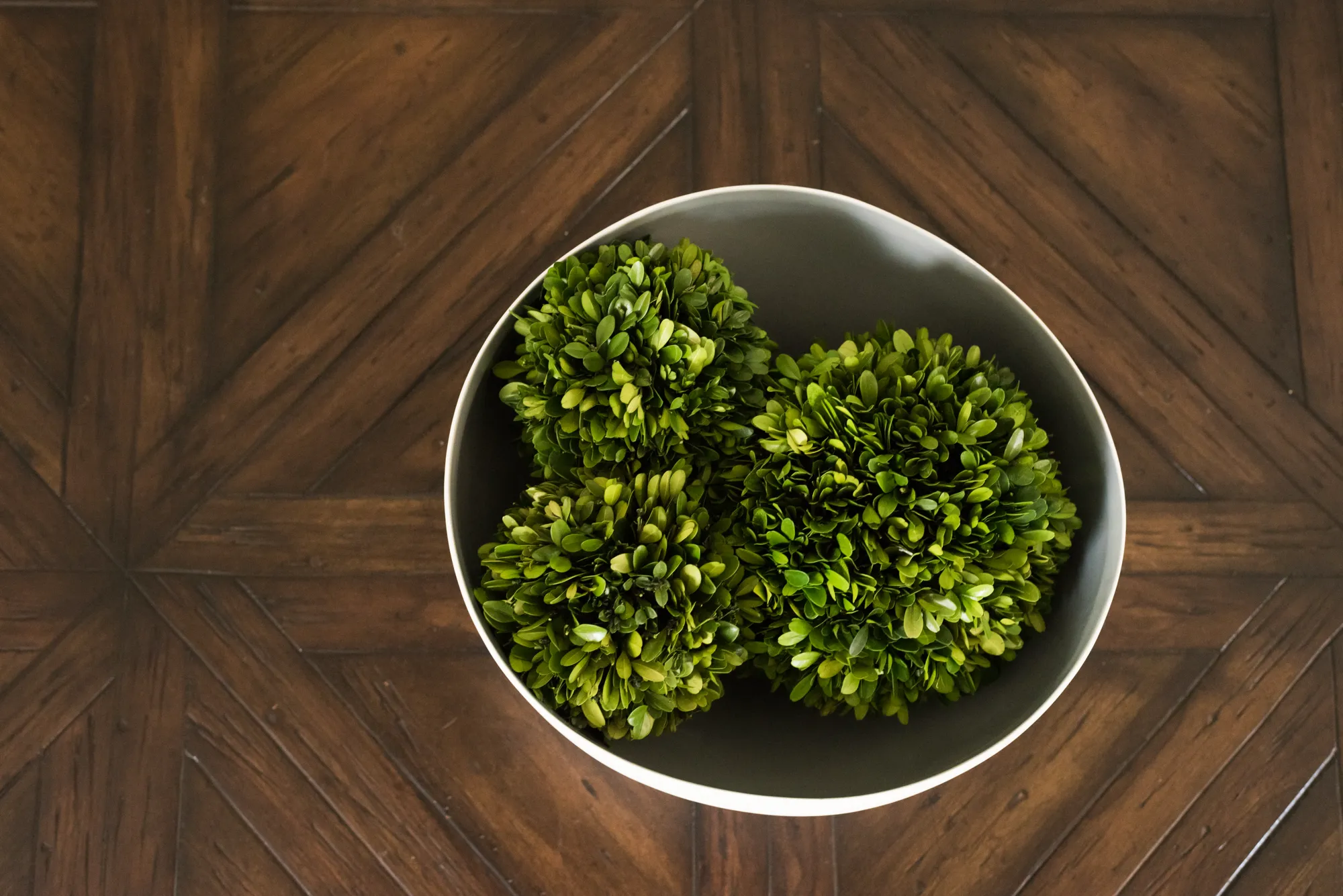 A neutral gray bowl holding decorative green boxwood balls rests on a dark wooden table with a parquet design.