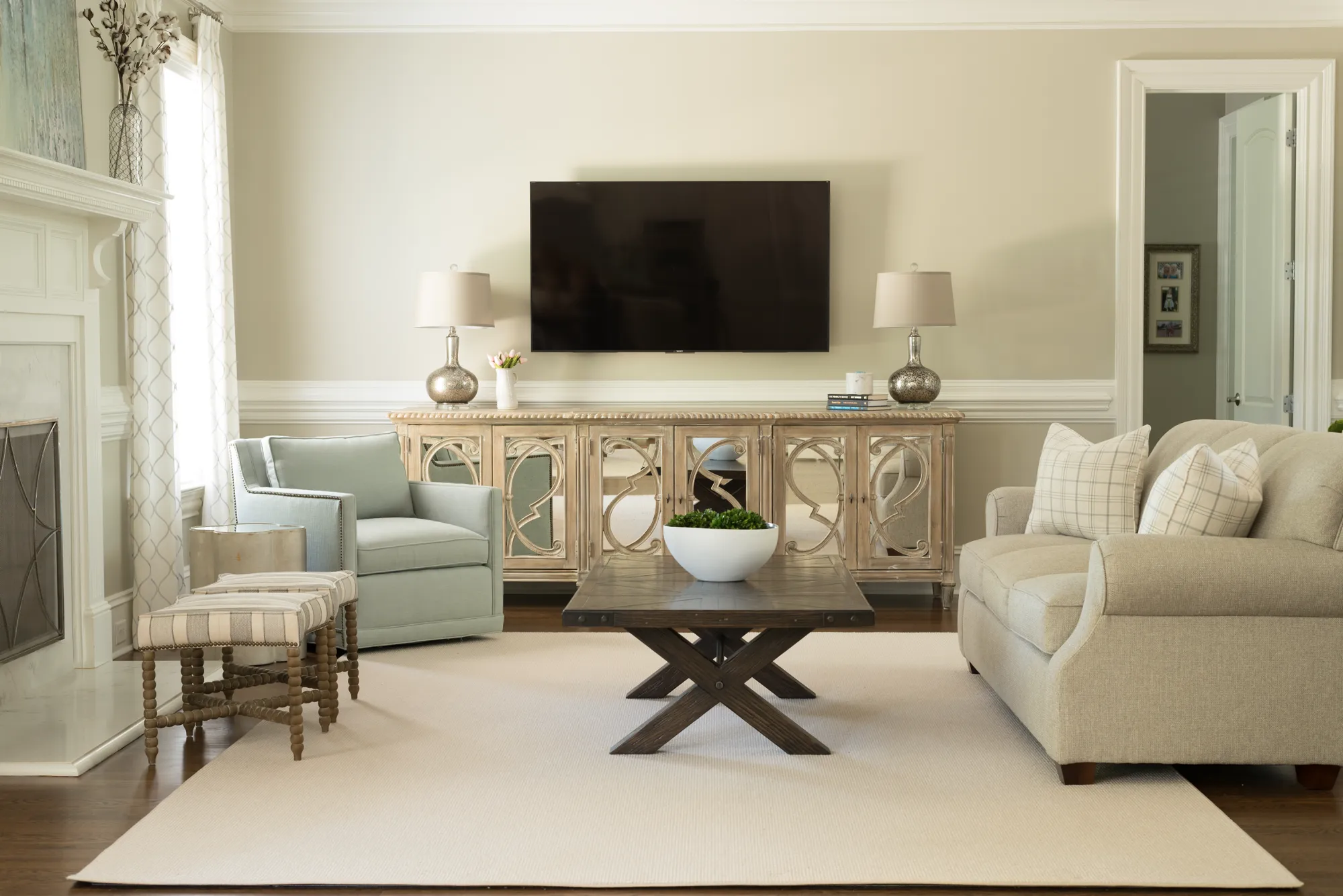 A sophisticated neutral living room featuring a TV mounted above a mirrored console, flanked by a light beige sofa and a light blue accent armchair.