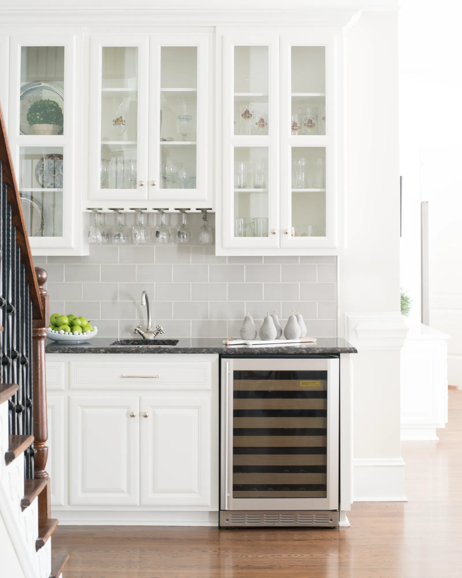 A home wet bar area, positioned next to a dark wood staircase, features white cabinets, a gray tile backsplash, and a built-in wine refrigerator.