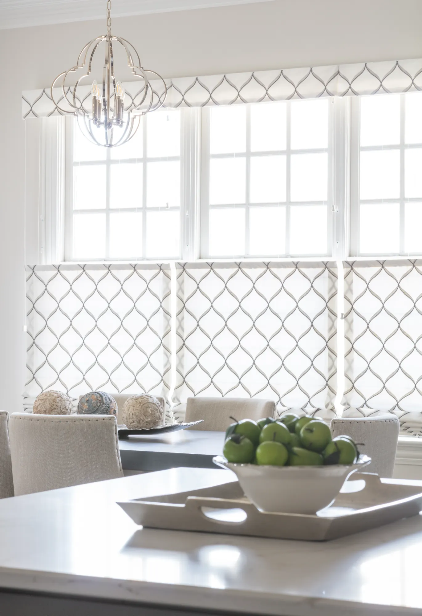 A dining room with geometric window shades and a metallic chandelier. A bowl of green apples rests on a white countertop in the foreground.