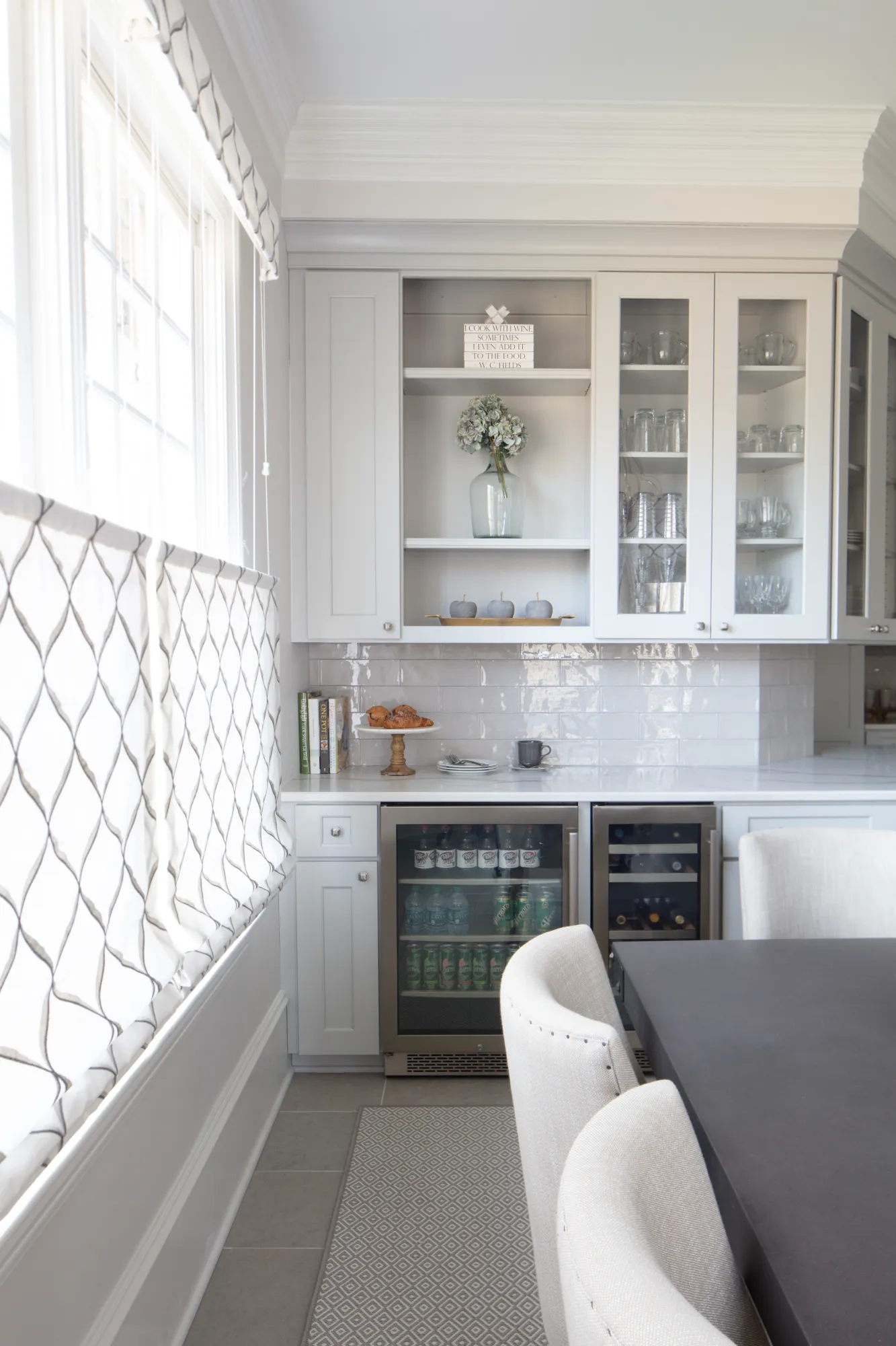 A brightly lit modern kitchen nook with white cabinetry, glass display shelves, two built-in beverage coolers, and a counter set with a light breakfast.