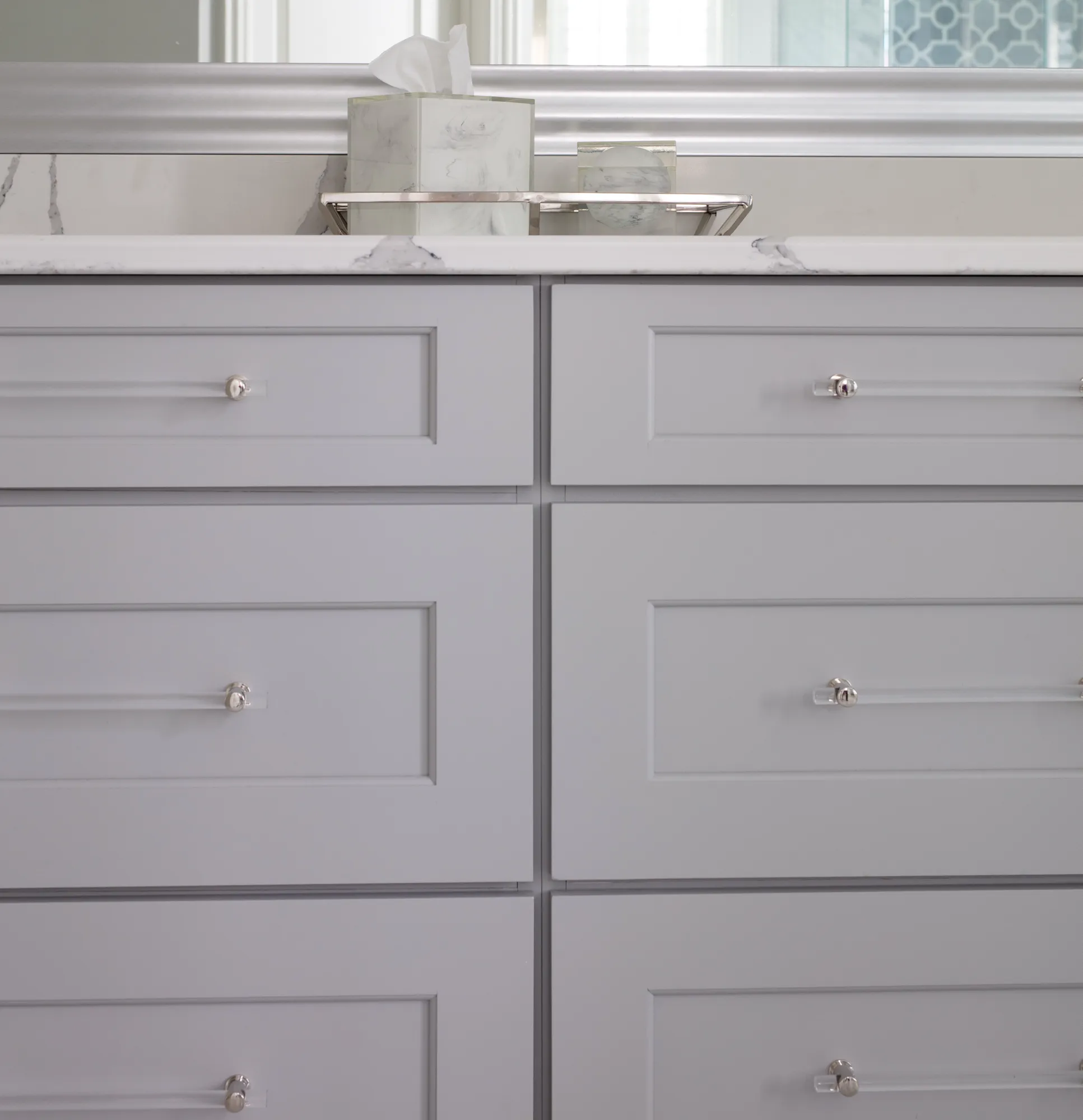 A modern, light gray shaker vanity with clear acrylic bar pulls. The white marble counter holds a decorative tissue box and accessories.