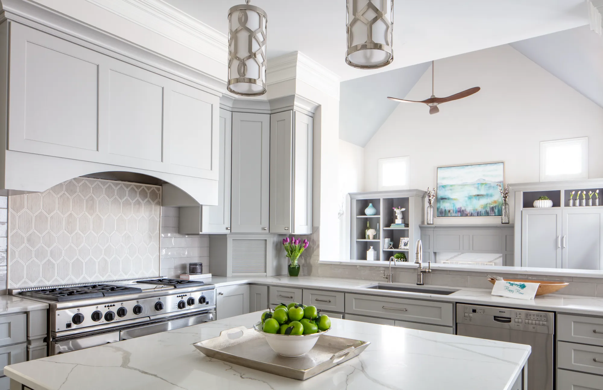 A bright, modern kitchen with light gray shaker cabinets, white marble countertops, and a hexagonal tile backsplash. Green apples sit on the central island.