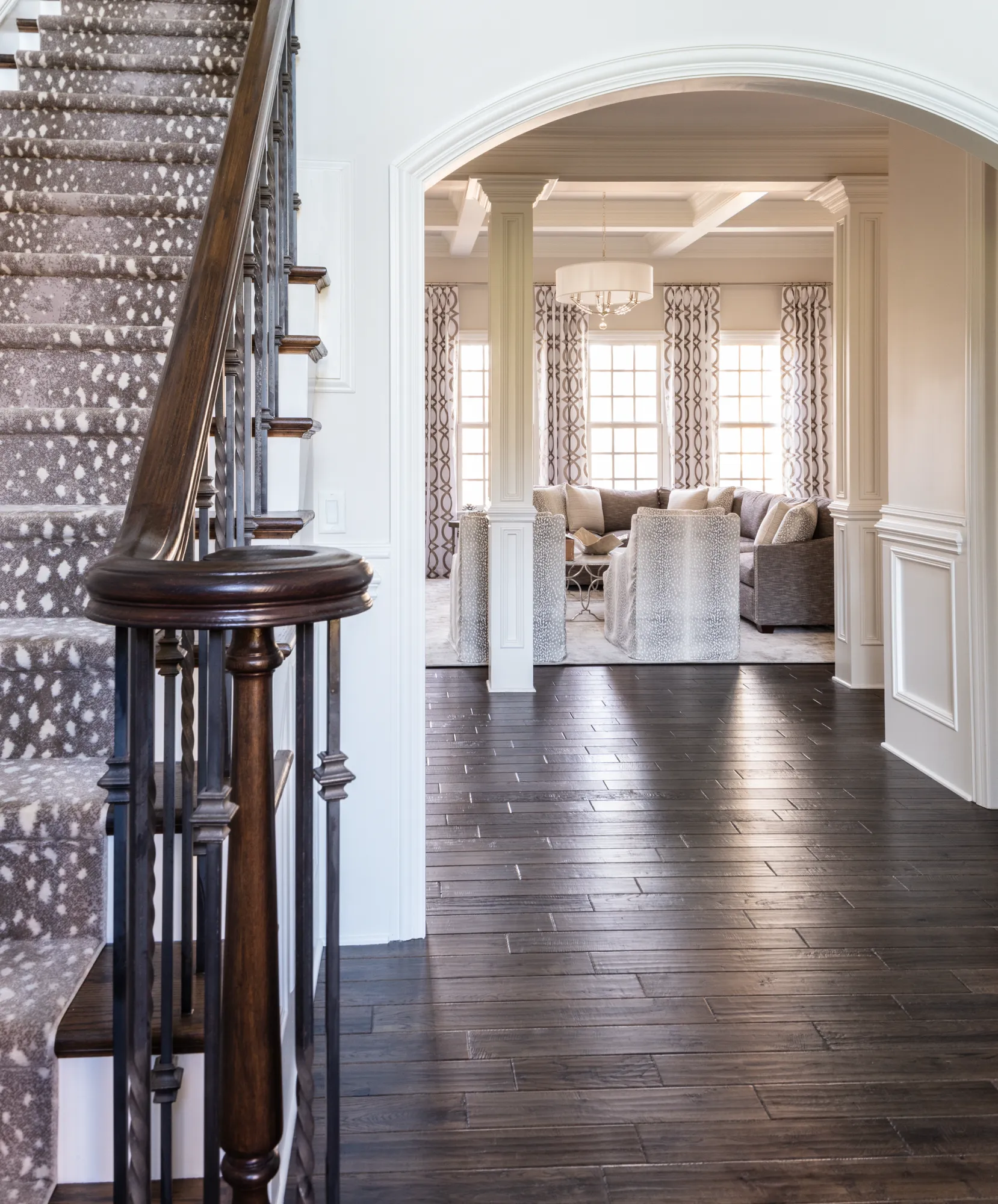 A well-lit entryway shows dark hardwood floors and a staircase with a spotted runner. An arch opens to a living room with a sectional and patterned accent chairs.