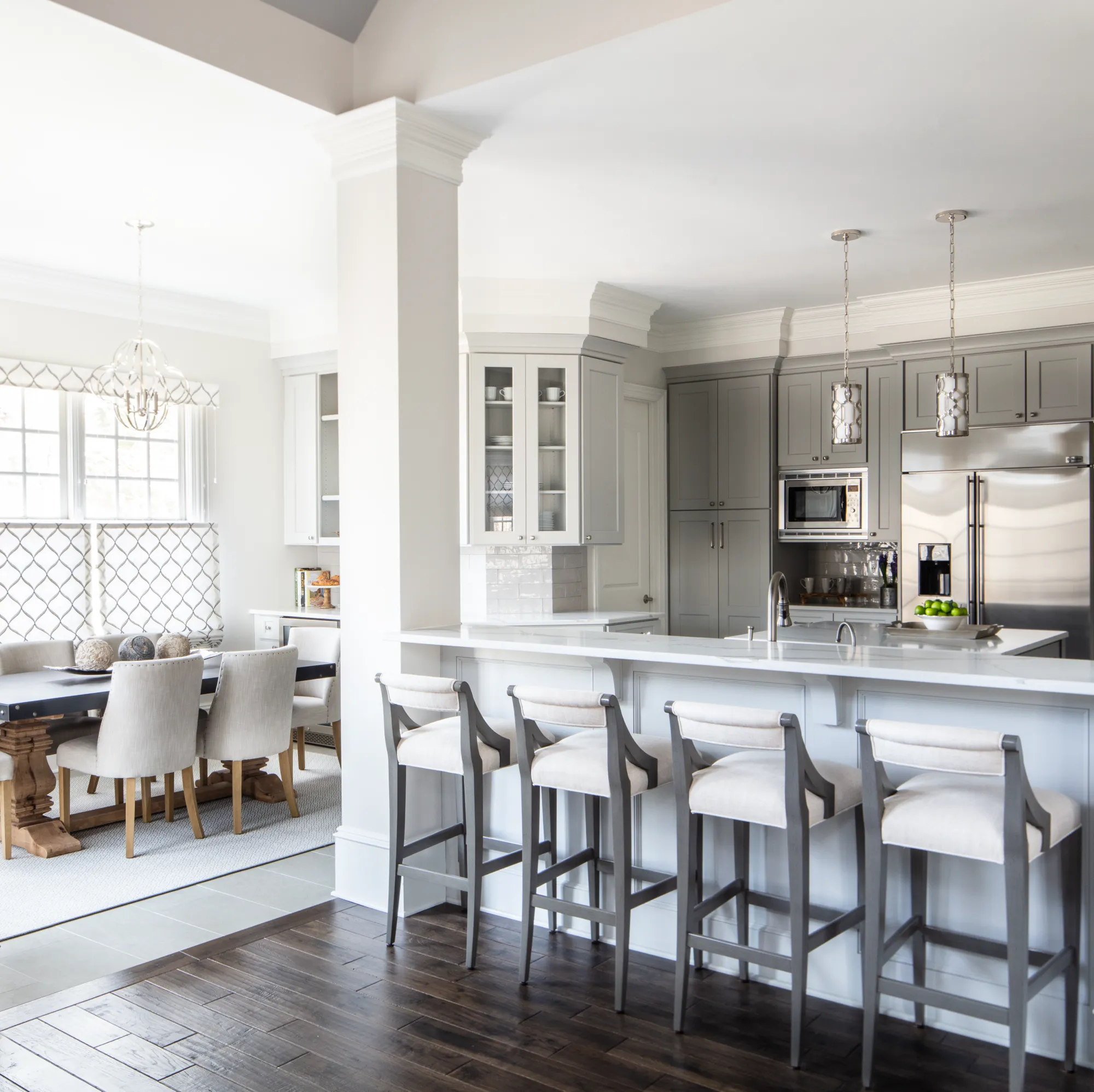 An open concept kitchen featuring gray cabinets and a white island with four neutral bar stools. The view extends to a bright dining area with a wooden table and upholstered chairs.