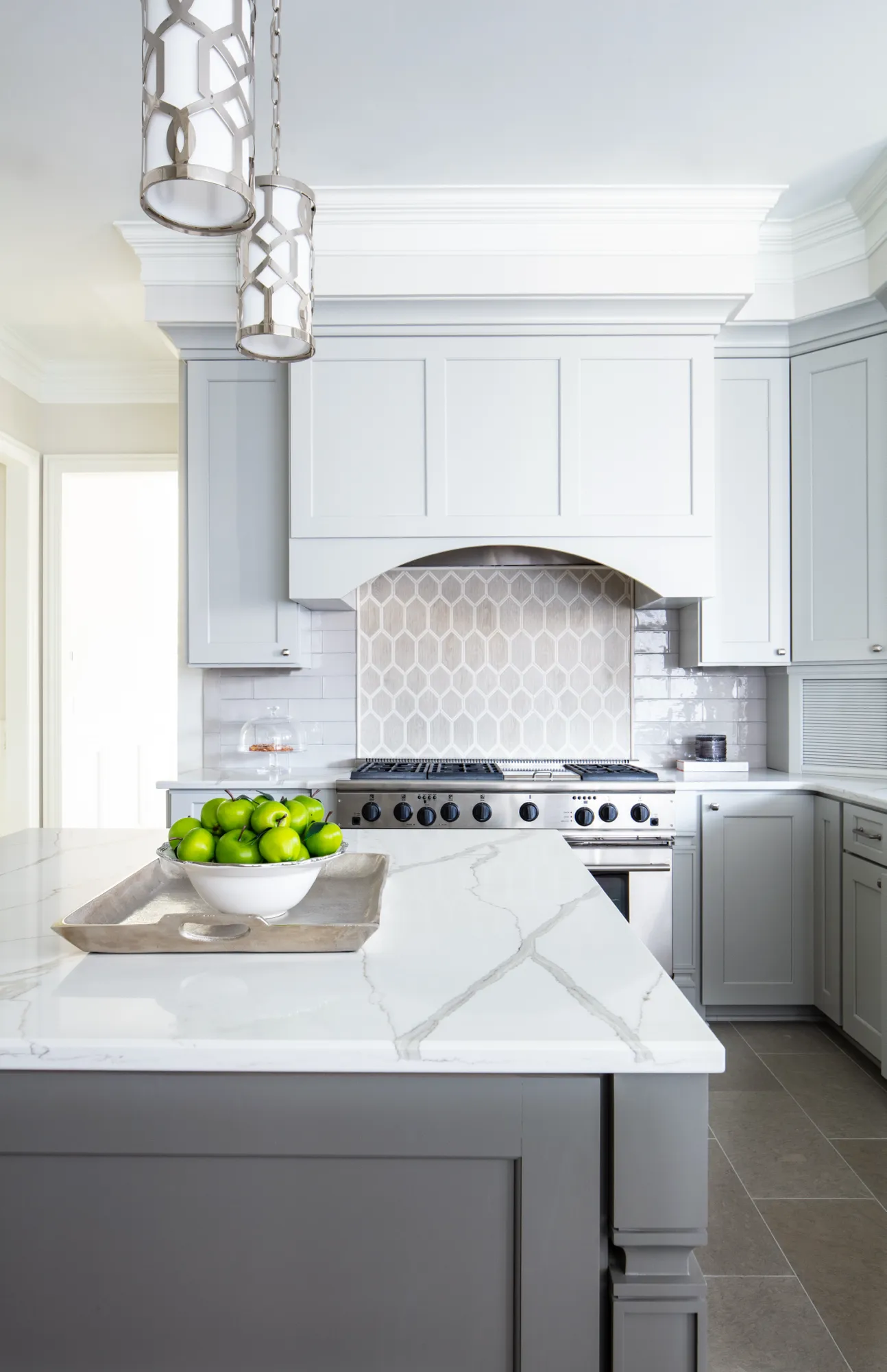A bright, modern kitchen featuring light gray cabinetry, a stainless steel range, and an elegant geometric backsplash. A bowl of green apples sits on the marble-top island.