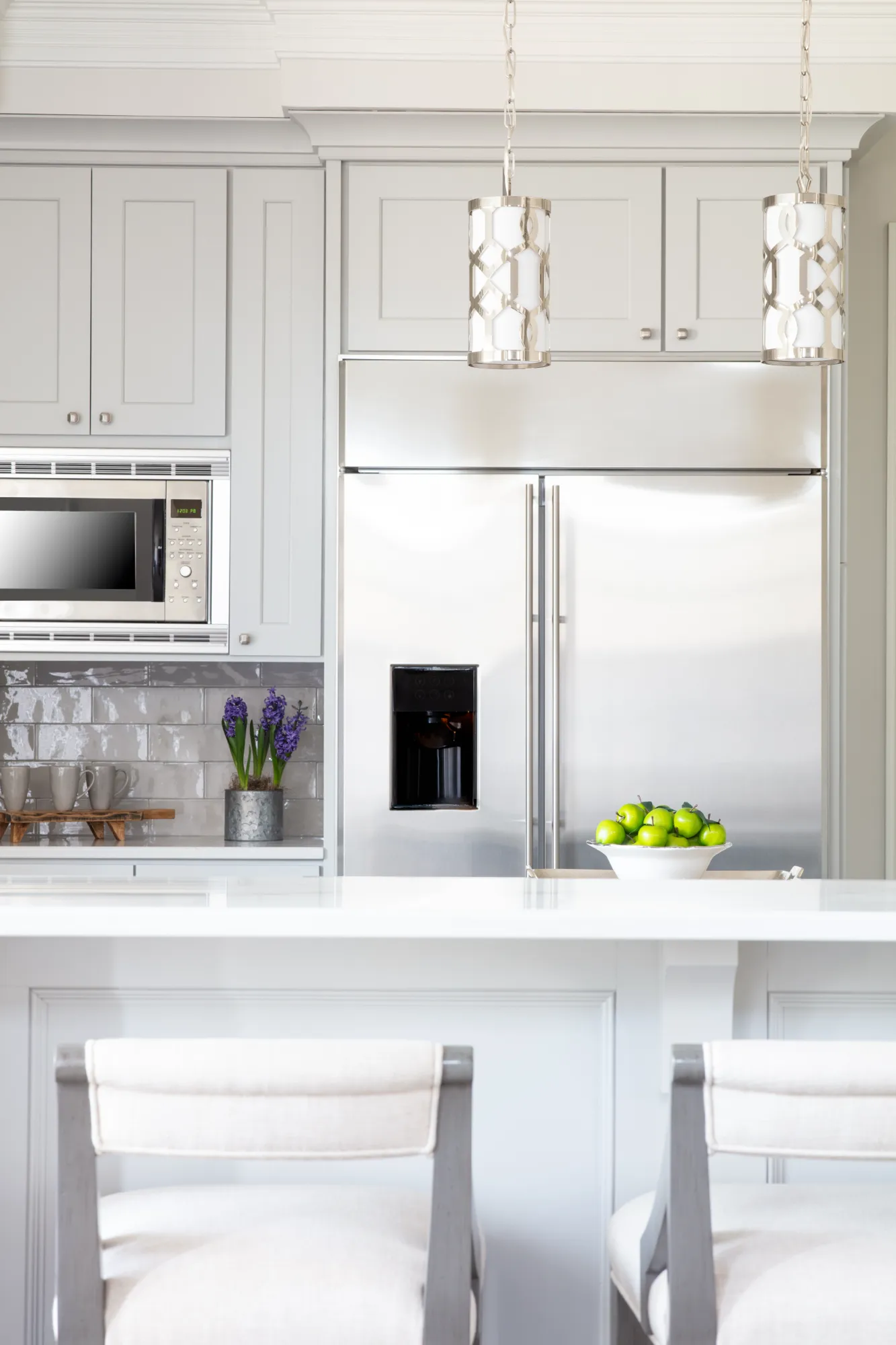 A sleek, light gray kitchen with a stainless steel refrigerator and microwave built into the cabinetry. A white island counter features two upholstered stools and green apples.