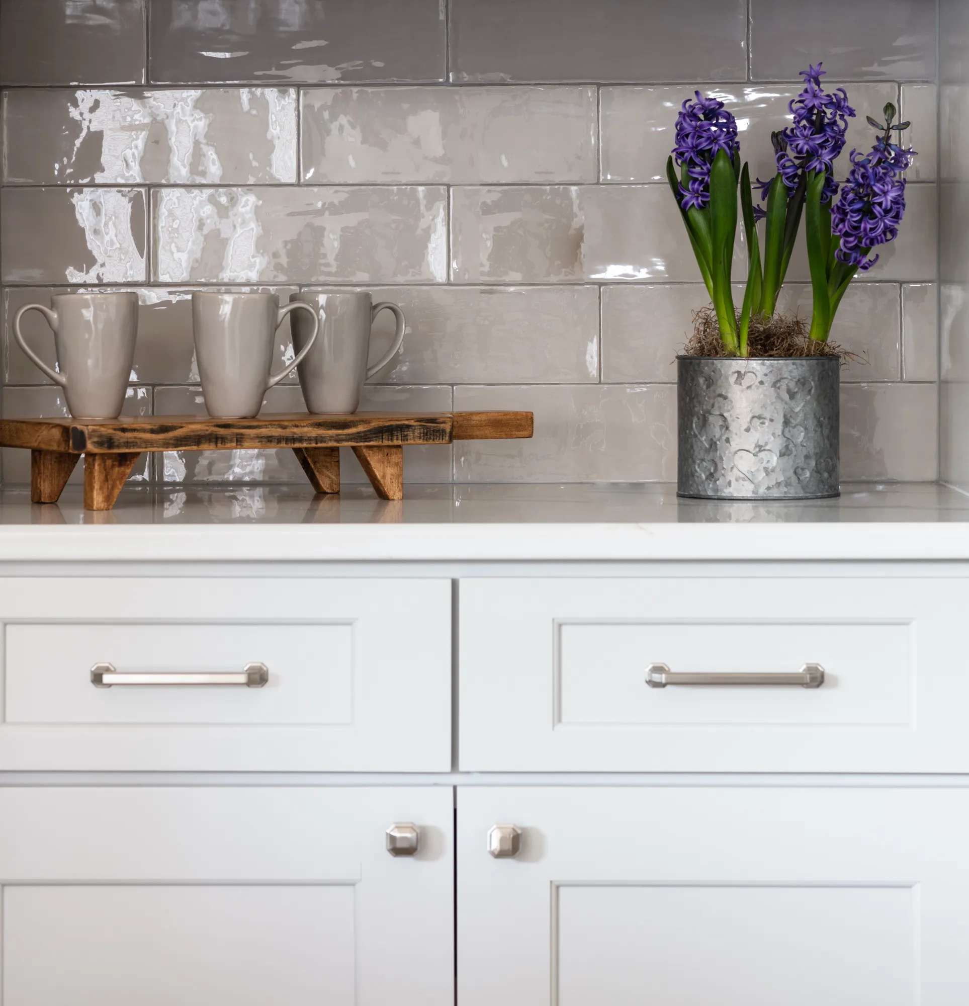 White kitchen cabinets and a glossy grey subway tile backsplash are decorated with three grey mugs on a wooden stand and vibrant purple hyacinth flowers.