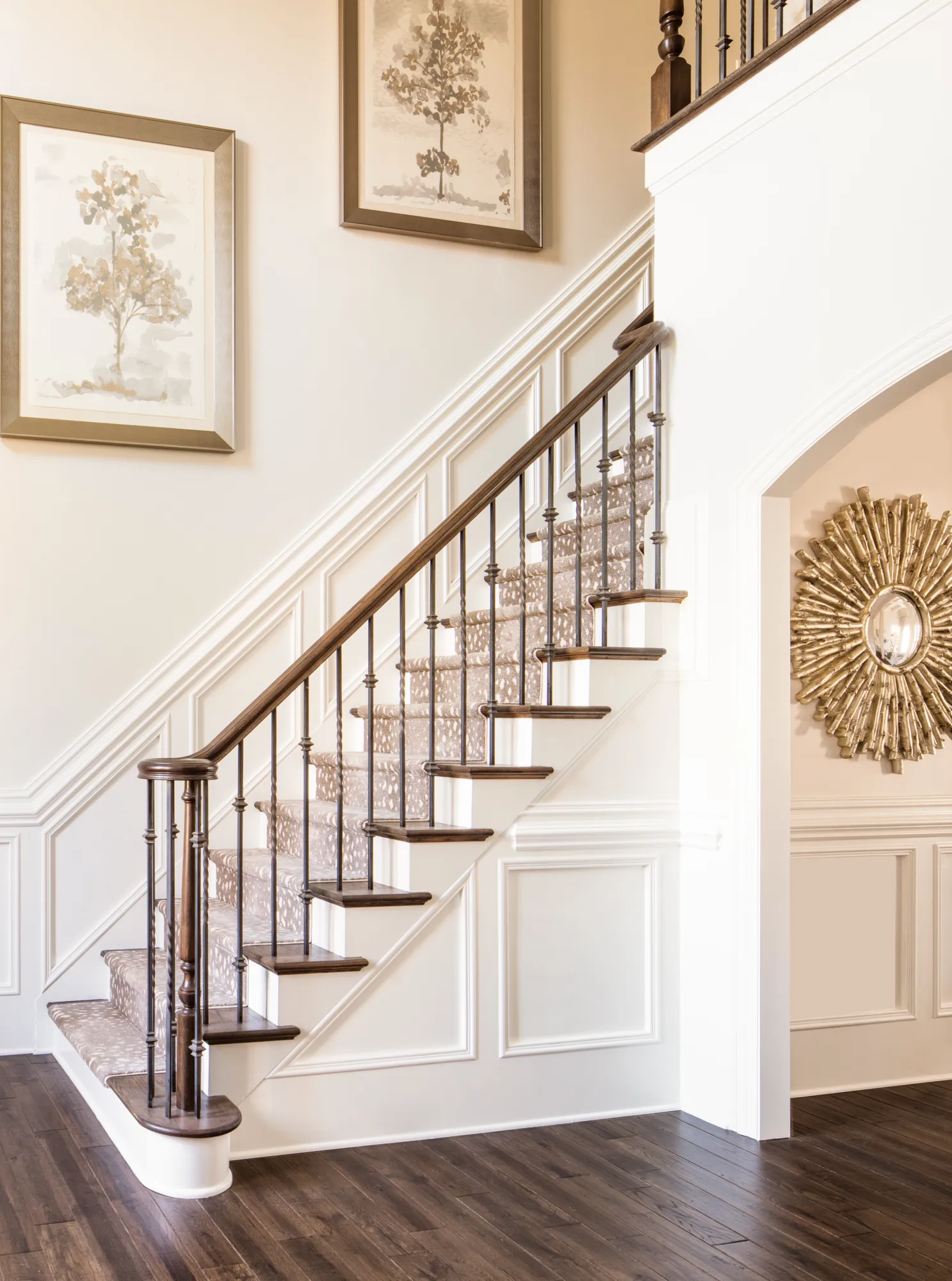 An elegant foyer featuring a curved staircase with a patterned runner, dark wood treads, and black iron balusters. The light walls have white wainscoting and two tree paintings.