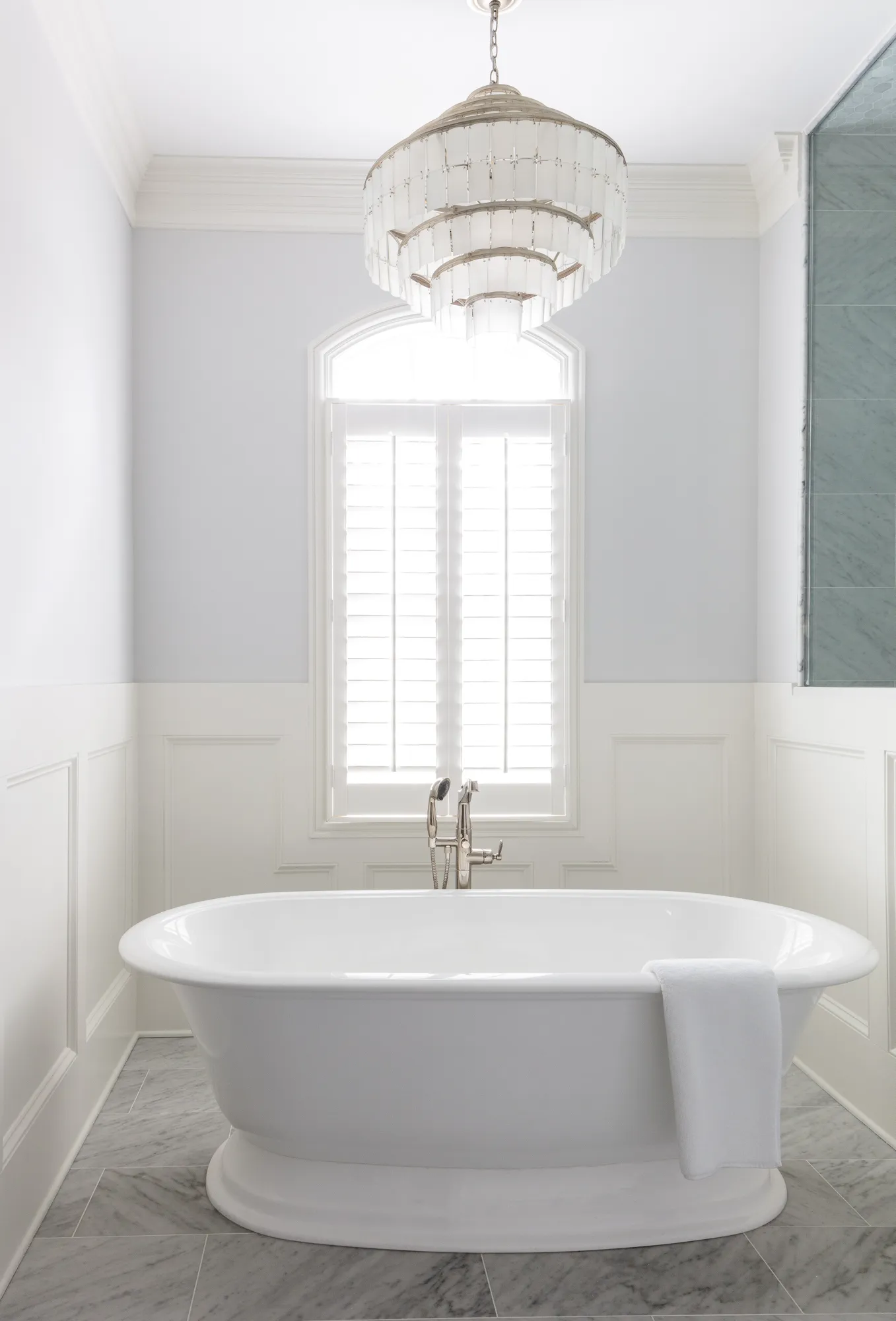 A white freestanding tub sits on gray marble tiles in a bright bathroom. Above the tub is an arched window with shutters and a tiered glass chandelier.