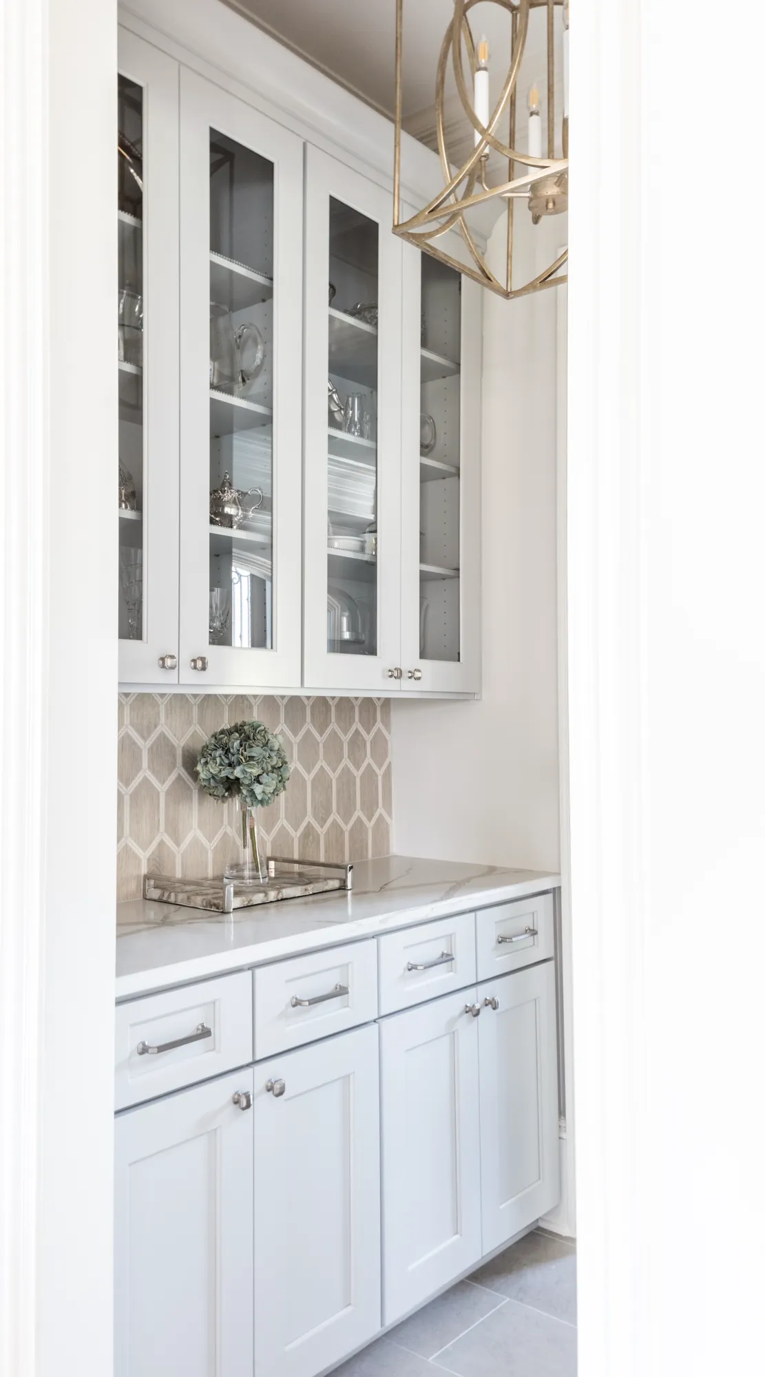 A bright butler’s pantry with light gray cabinets, a hexagonal backsplash, and glass-front uppers. A gold geometric chandelier illuminates the space.