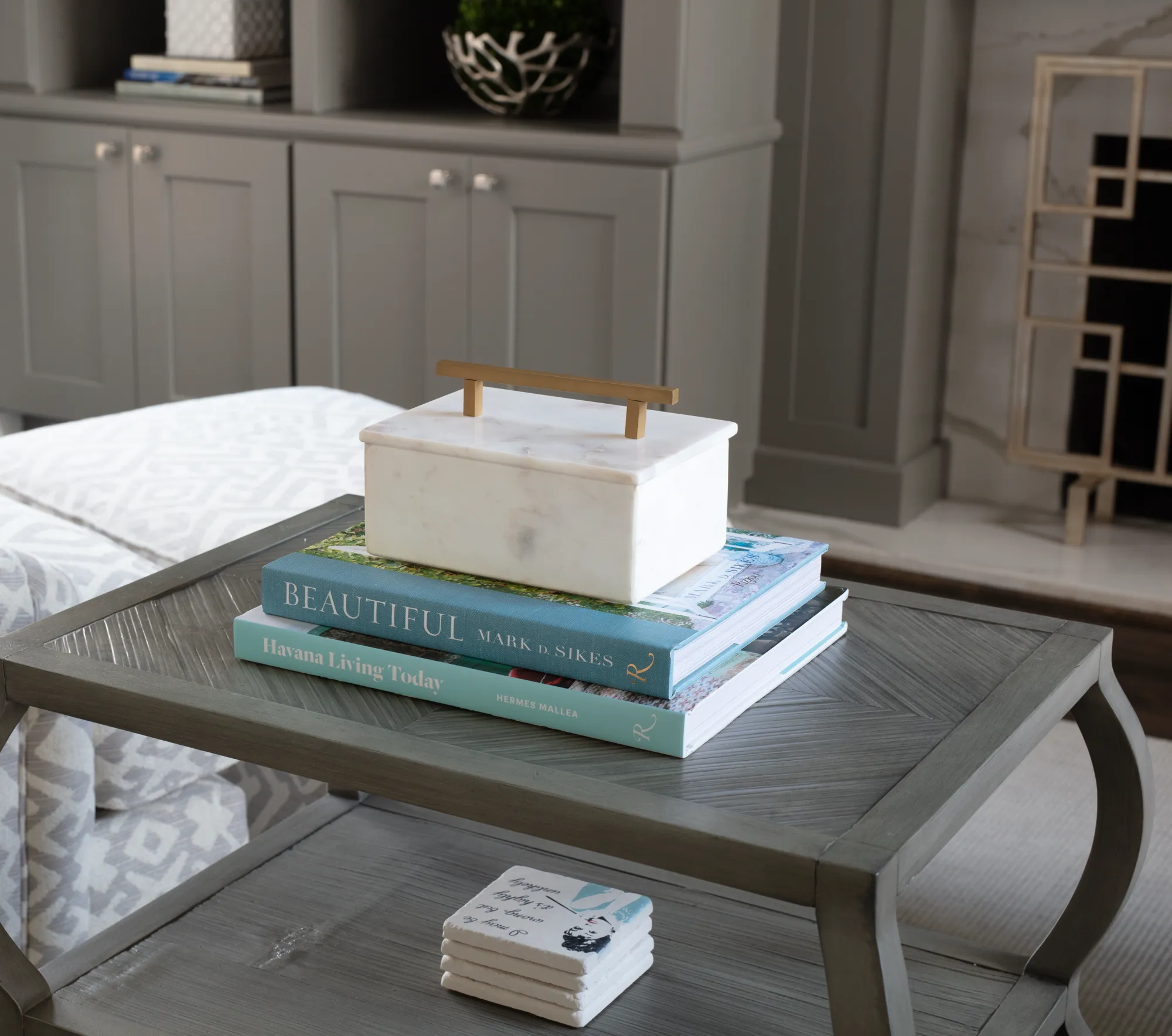 A gray accent table is styled with two coffee table books topped by a white marble box with a brass handle. A stack of coasters sits on the lower shelf.