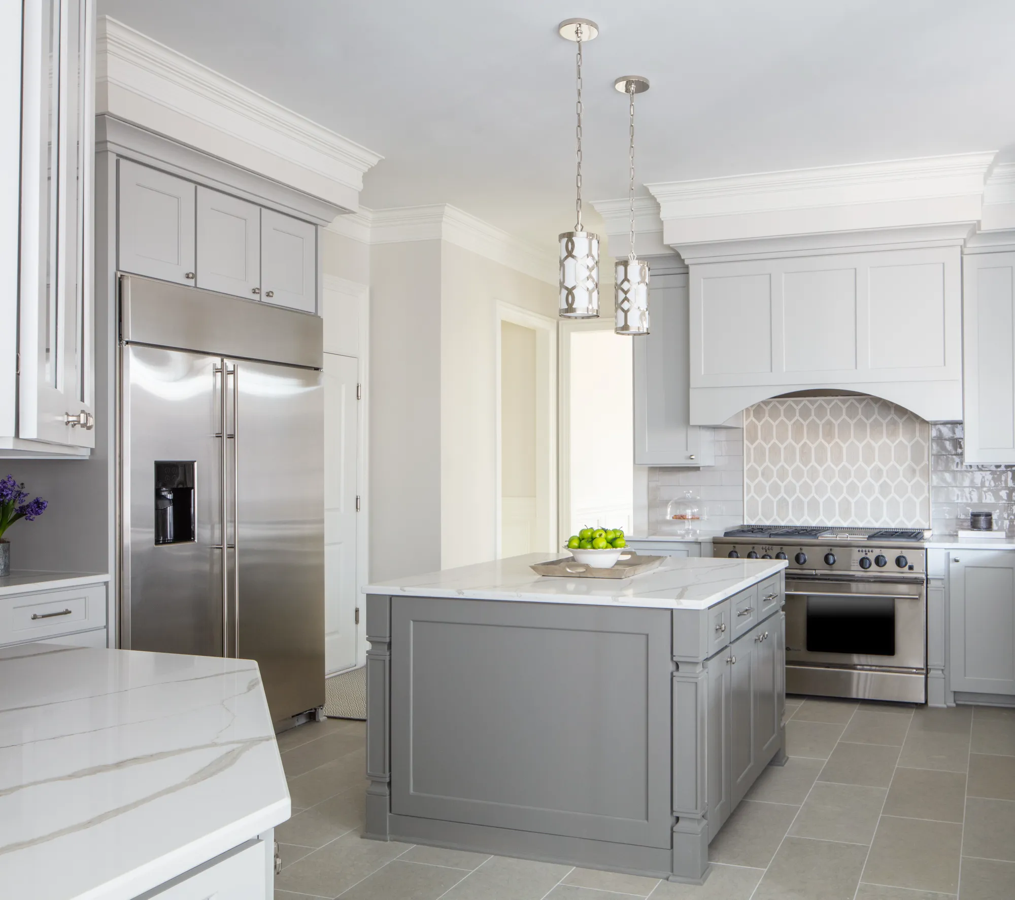 A contemporary kitchen with light gray cabinetry and a built-in stainless steel refrigerator. A gray center island features a white marble top and two hanging pendant lights. A patterned backsplash is visible above the professional range.