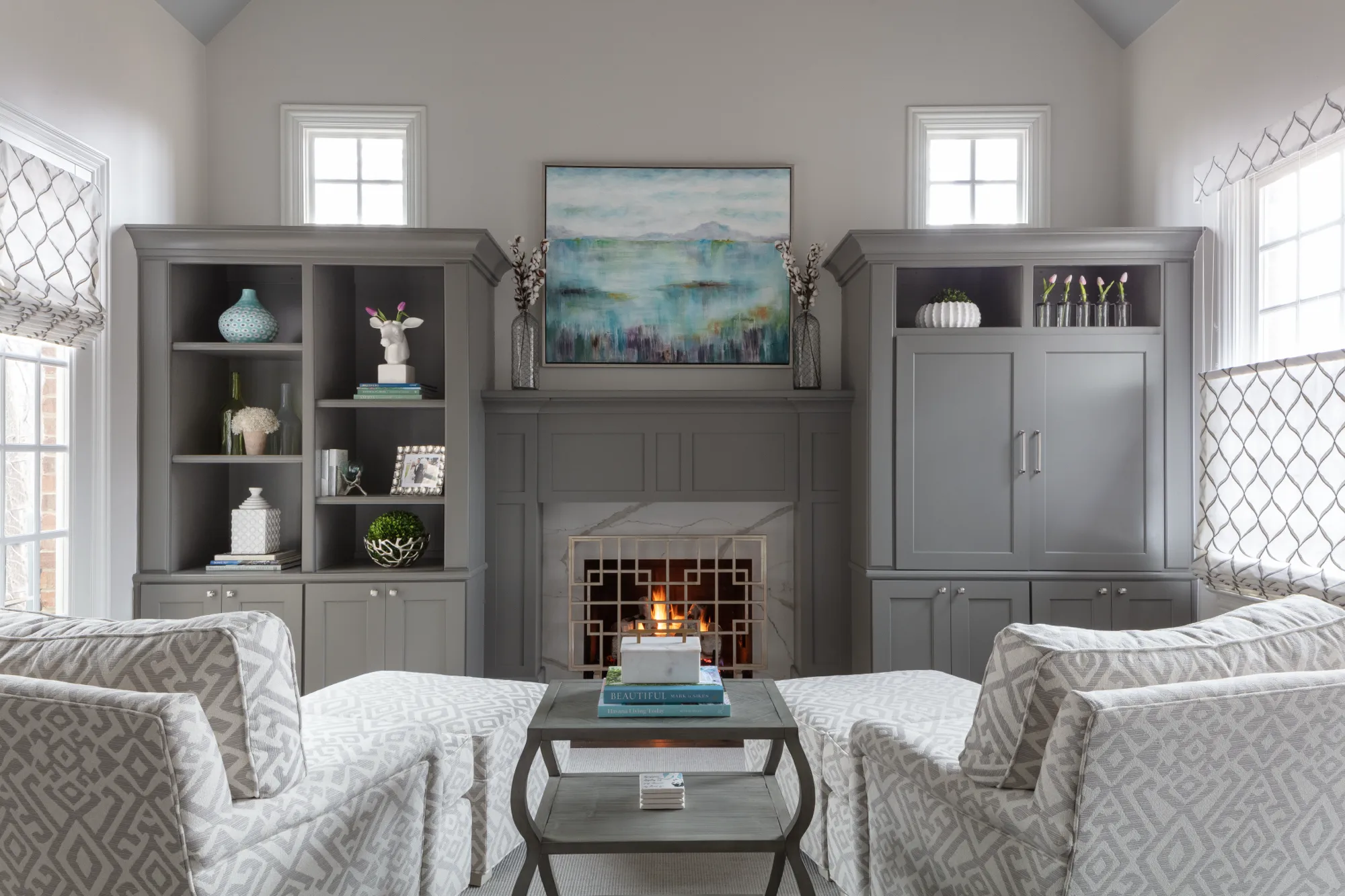 A symmetrical gray living room features a fireplace flanked by built-in cabinets, with a striking abstract painting above. A small center table is in between two patterned chaise lounges.