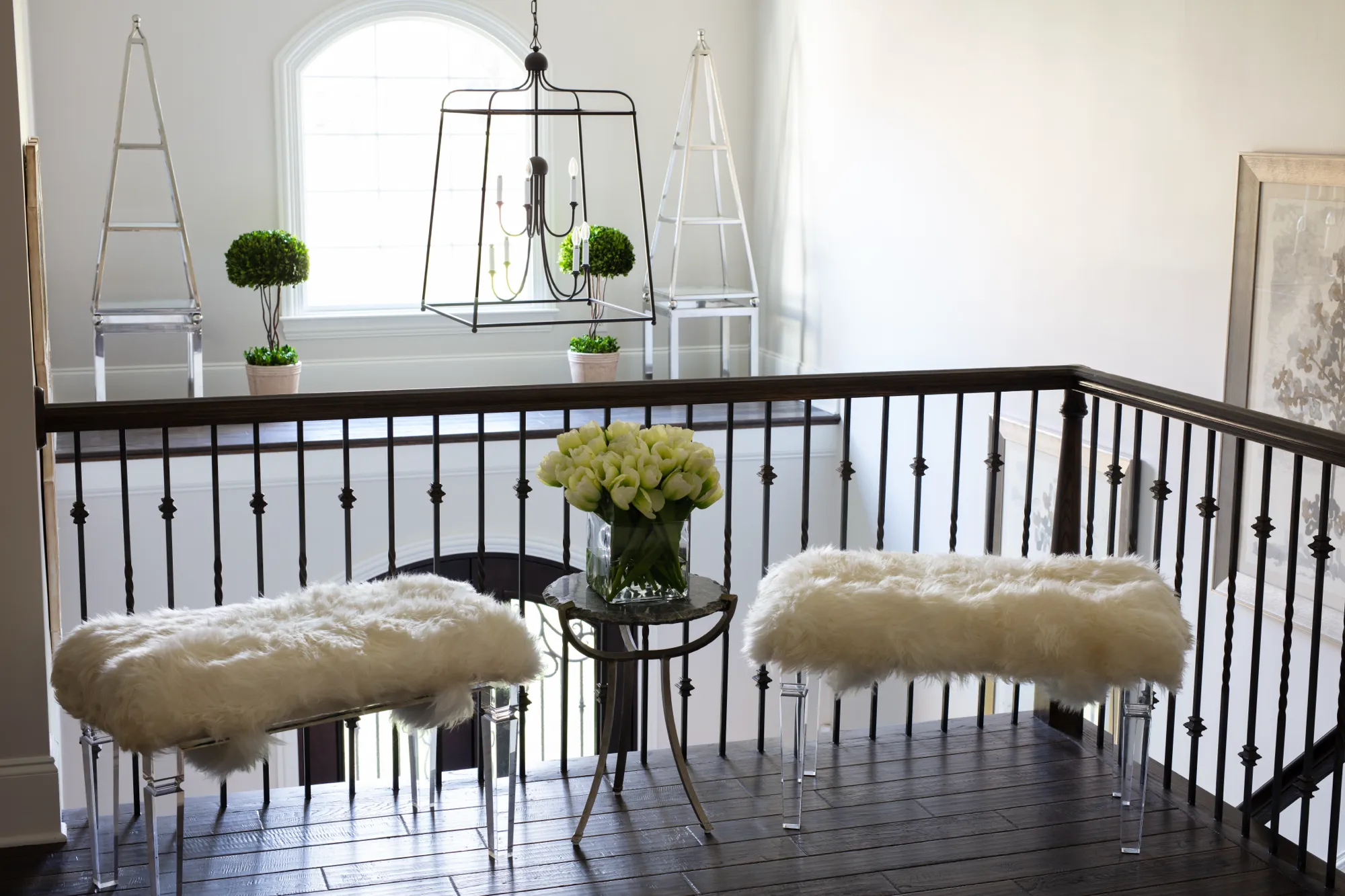 A chic interior design setup on a landing with dark wood floors. Two white fur benches flank a side table with white tulips, set against a black metal railing.