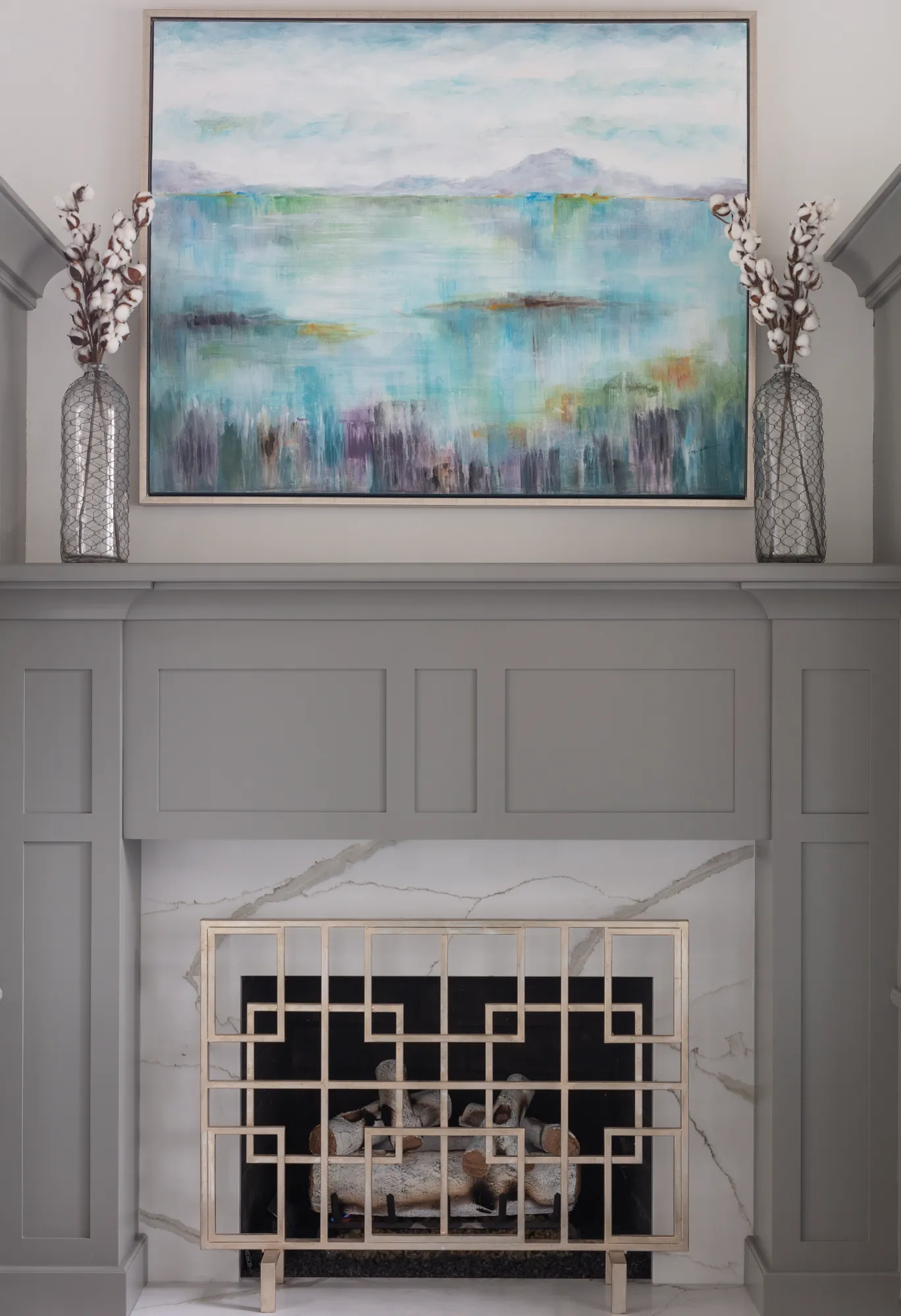 A gray paneled fireplace mantel topped by an abstract blue and green landscape painting, flanked by cotton branches in wire vases. The hearth features white veined marble and a gold geometric fire screen.