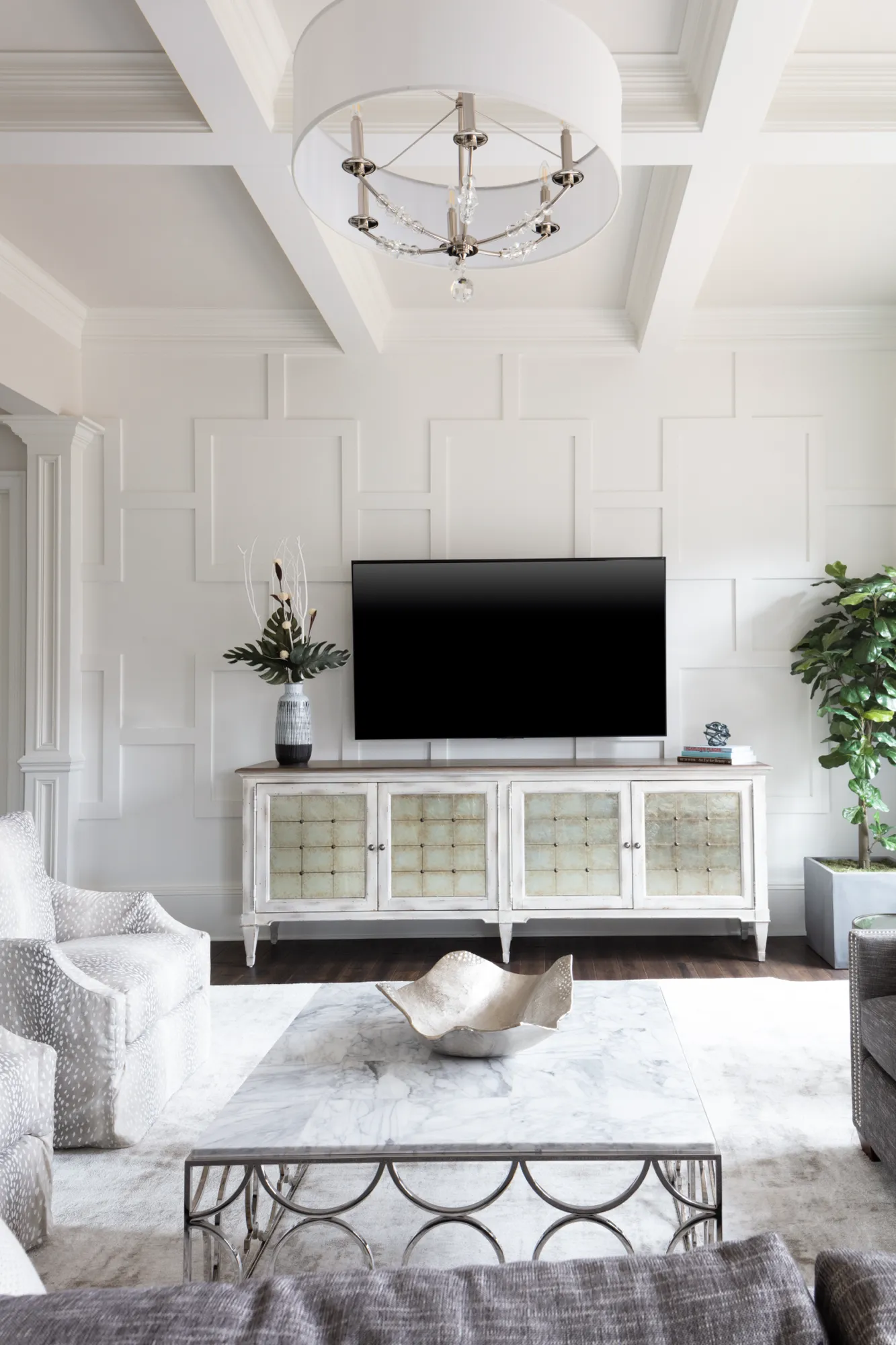 A contemporary white living room features decorative wall paneling, a coffered ceiling, and a large chandelier. A TV sits above a foggy-mirrored credenza, centered in front of a marble coffee table.