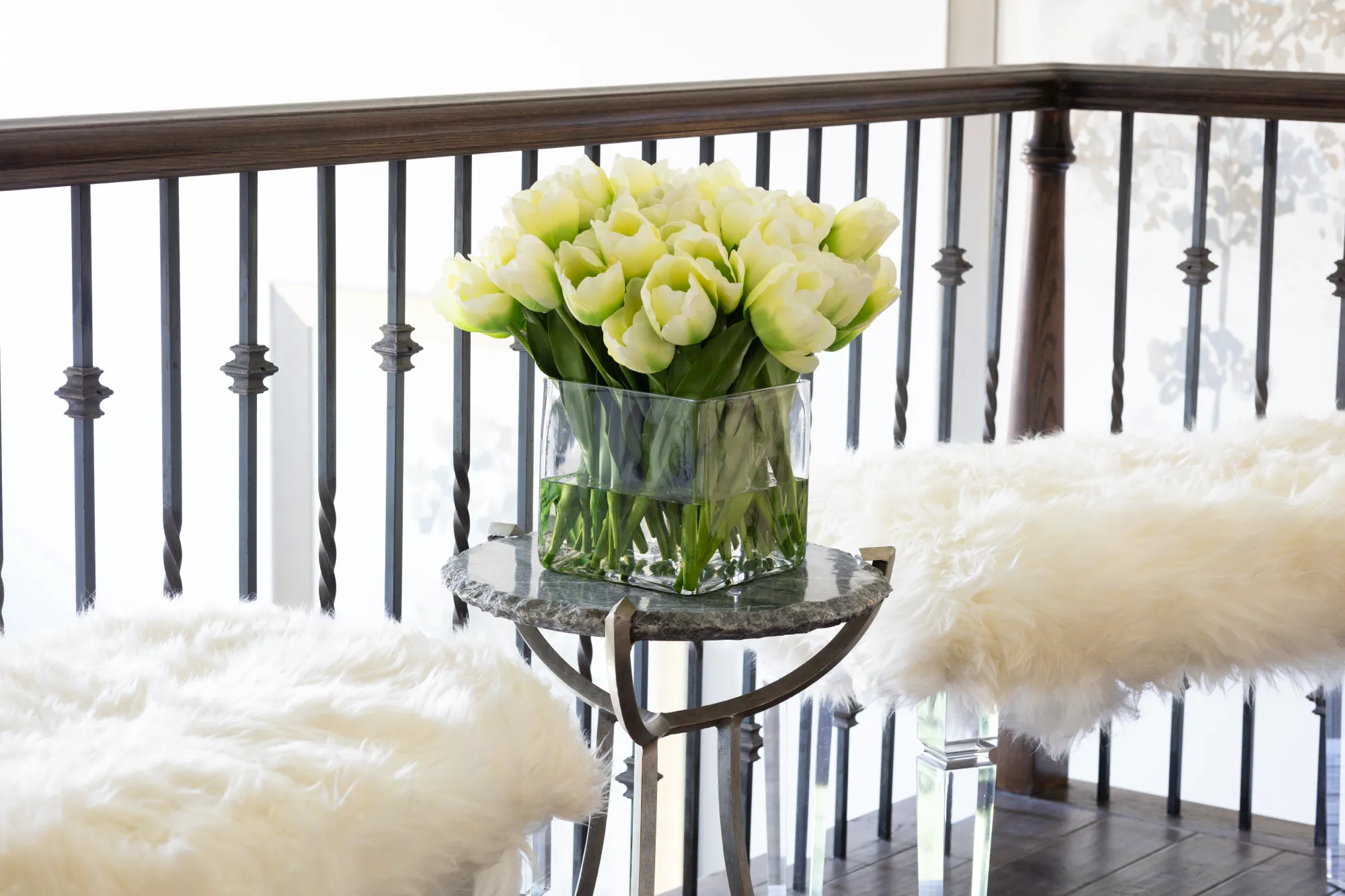 A large arrangement of cream and green tulips in a glass vase sits on a round stone side table. Plush white faux fur benches flank the table next to a dark wrought iron railing on a landing.