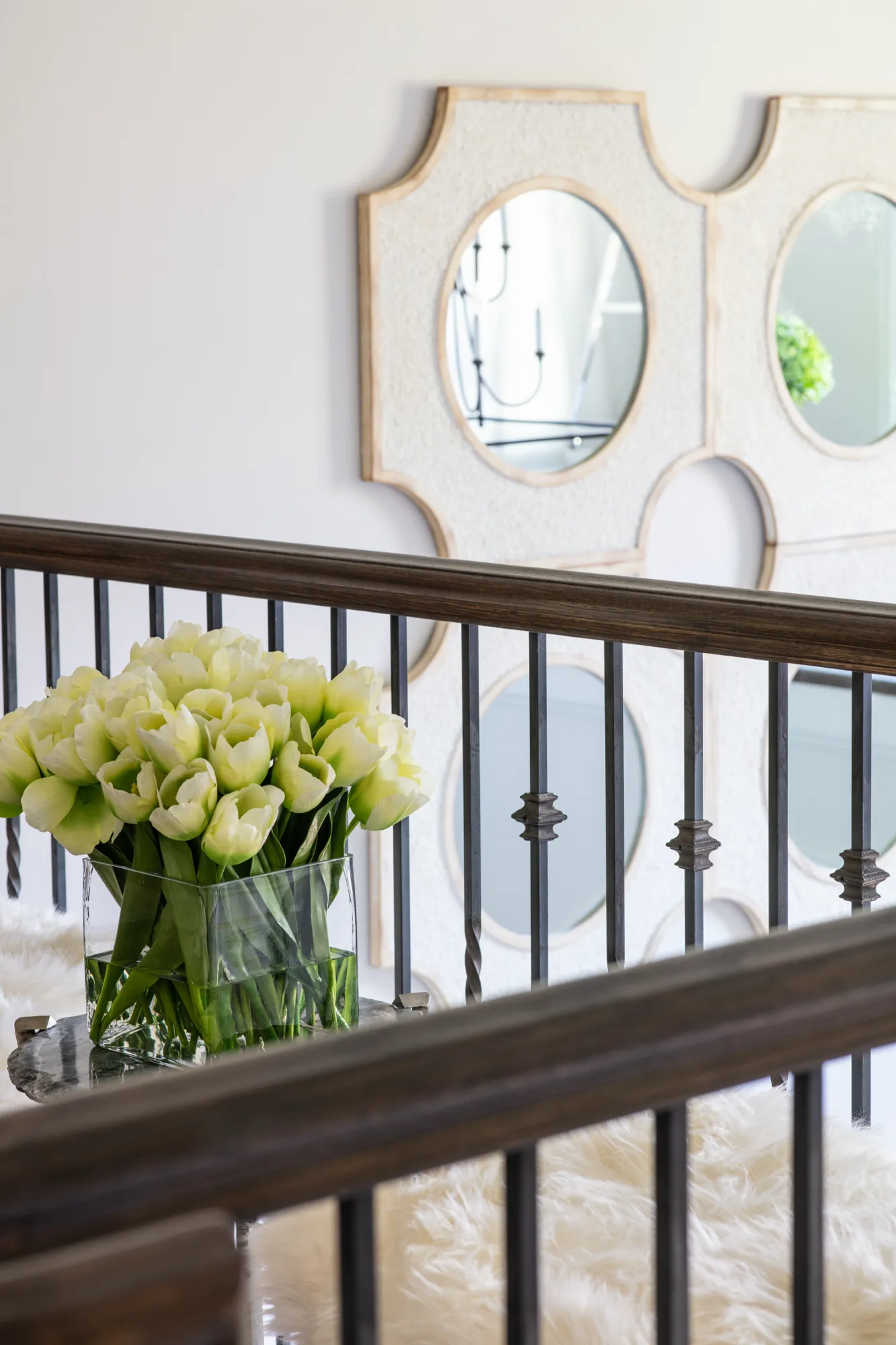A vase of pale green and white tulips rests on a stair landing. Dark wood railings frame the view toward decorative, ornate wall mirrors.