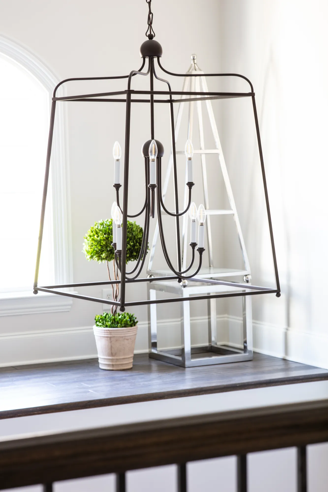 A contemporary, dark bronze lantern chandelier hangs above a small landing with a potted green plant and a mirrored display stand. Bright light enters from an arched window.
