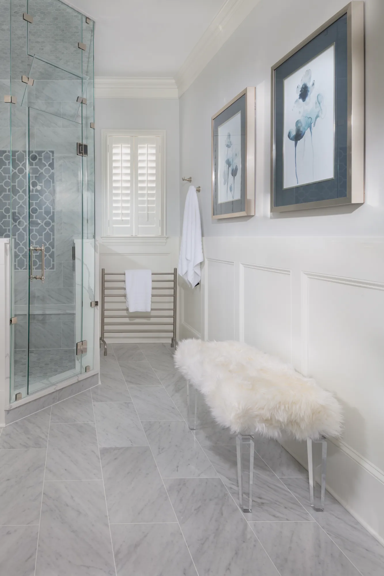 An elegant bathroom with gray marble tile flooring and a glass shower. The wainscoted wall holds two framed abstract blue artworks above a fluffy white bench.