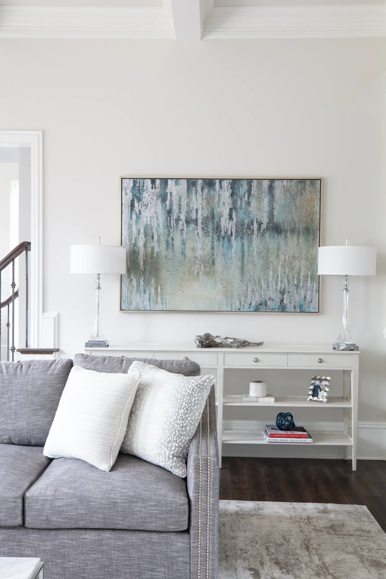 A white console table sits beneath a large abstract blue and gray painting, flanked by two tall crystal lamps in an elegantly decorated living room.