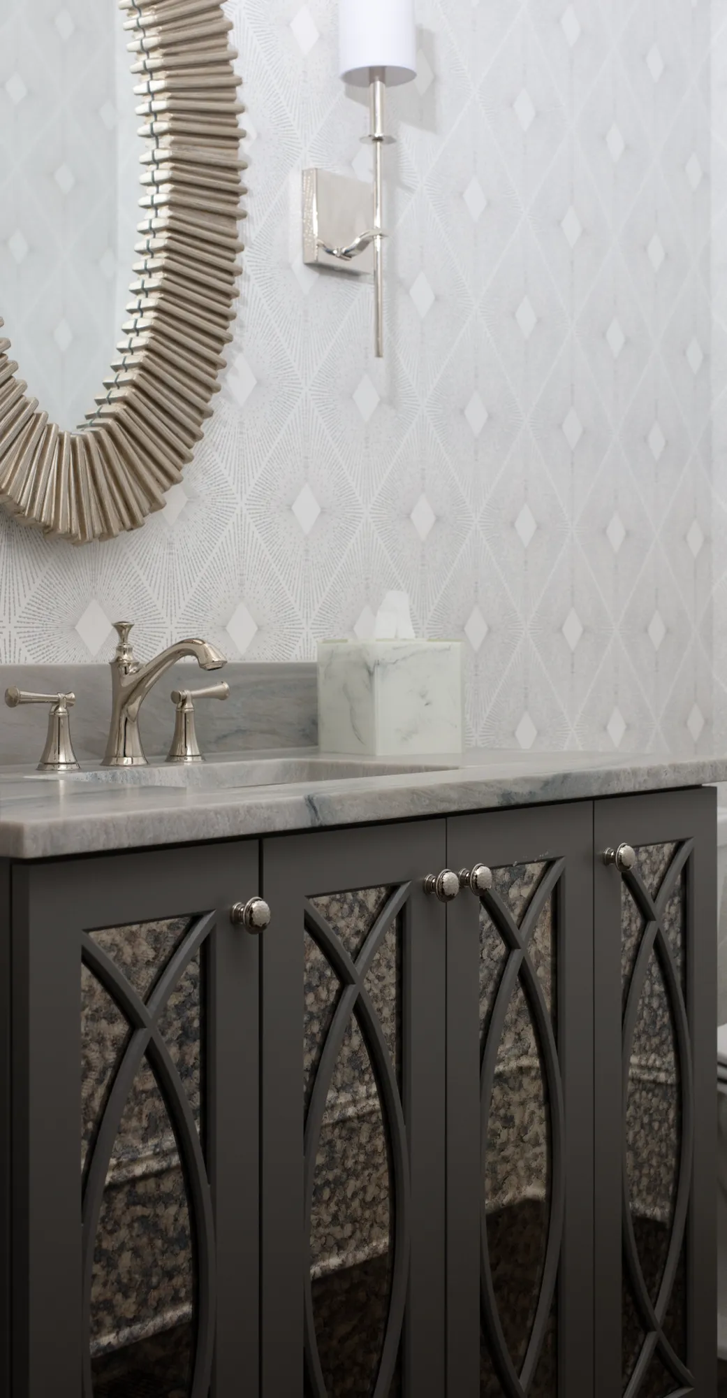 A dark gray bathroom vanity with decorative door panels and a marble counter holds a chrome faucet and a marble tissue box, set against patterned wallpaper.