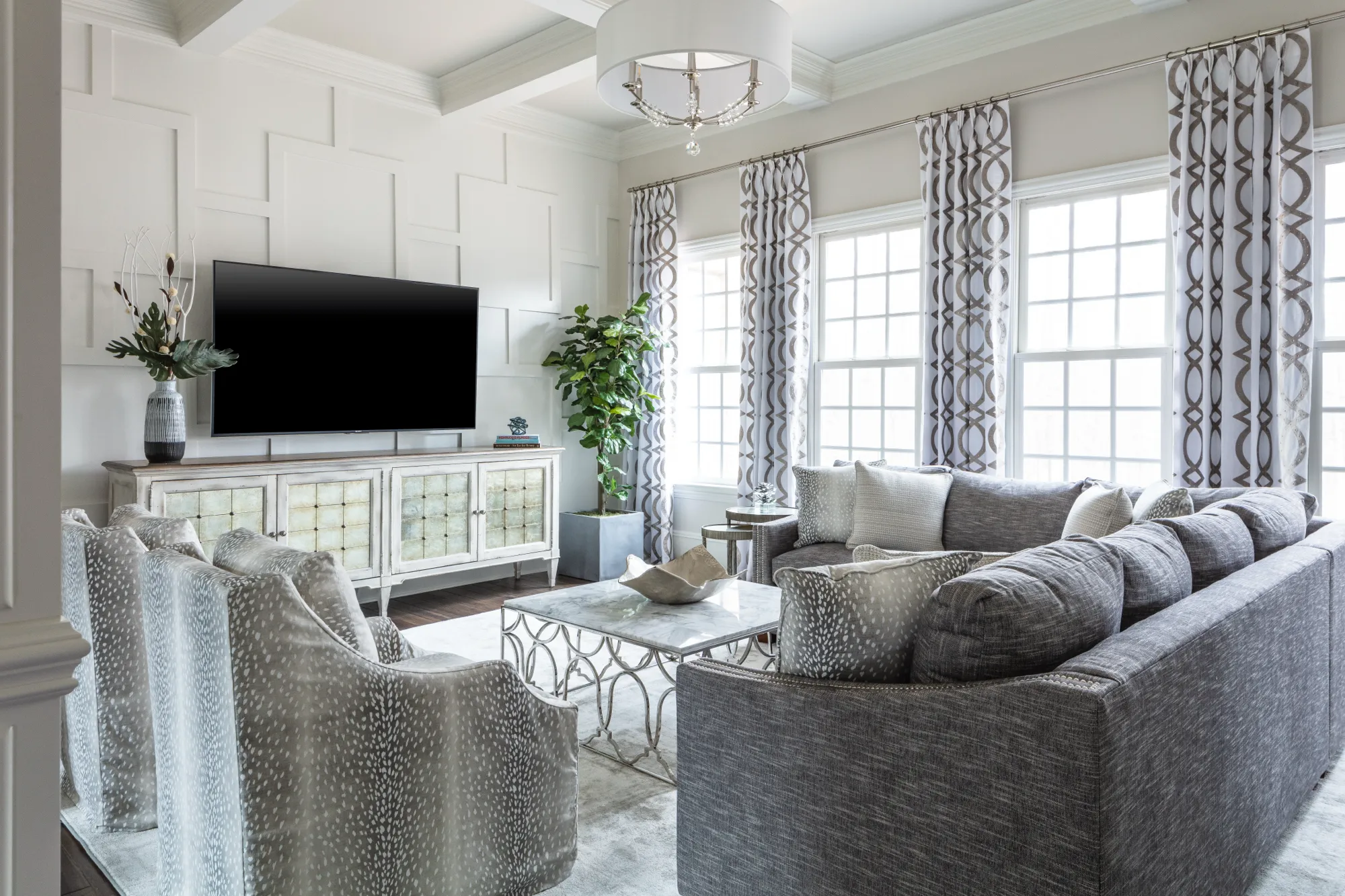 A sophisticated living room featuring white geometric wainscoting and a beamed ceiling. Gray textured seating, patterned curtains, and a marble coffee table fill the bright space.