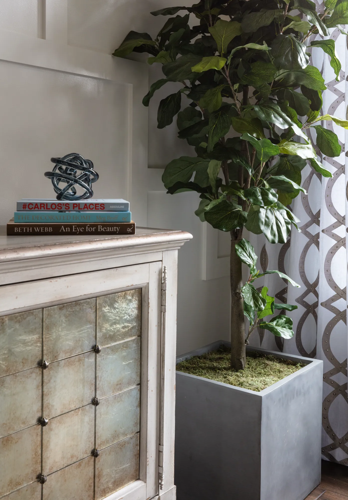 An interior vignette featuring a light-colored cabinet with decorative glass panels. Next to it stands a large potted tree in a square gray planter, set against a white patterned curtain.