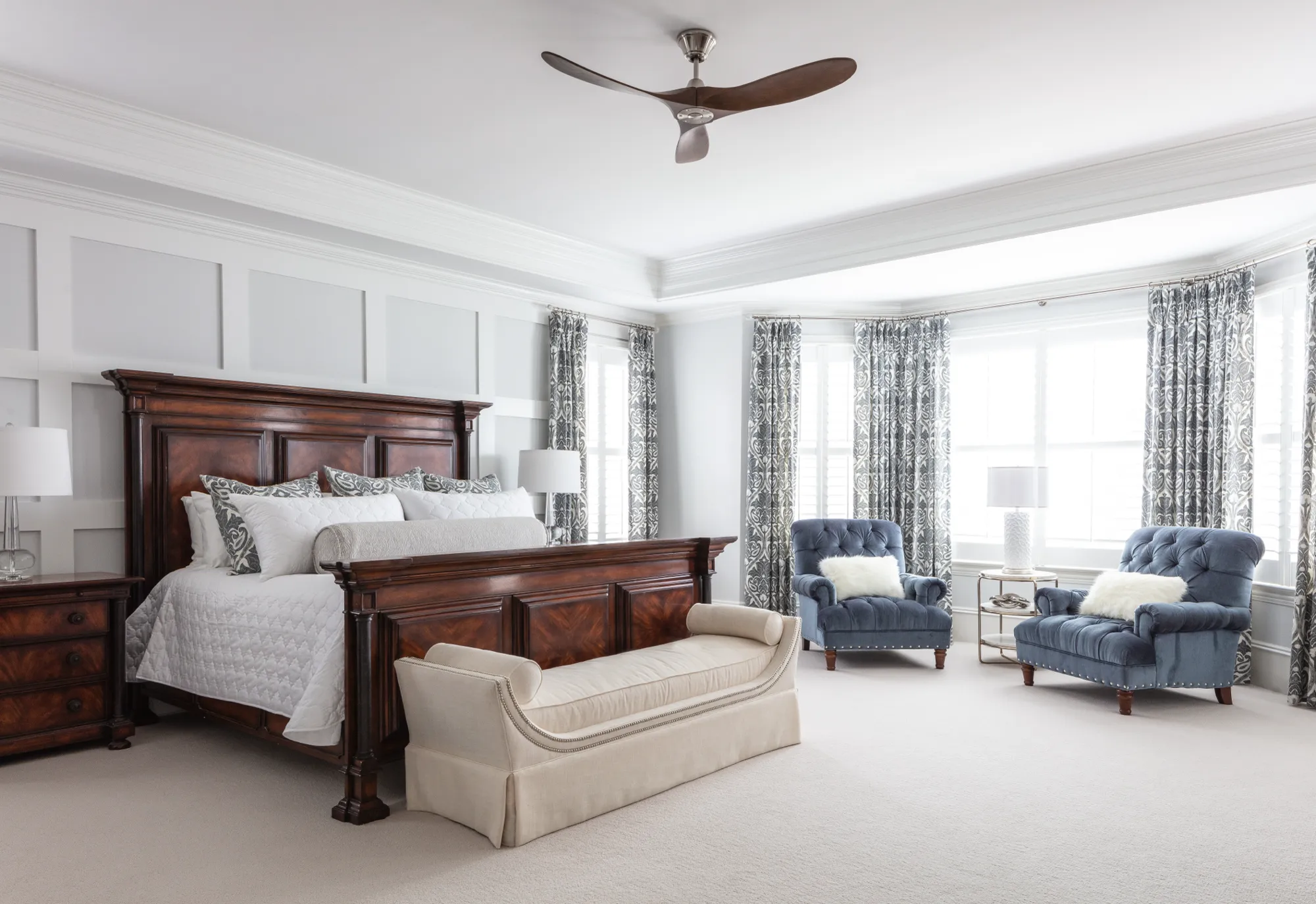A large bedroom with a dark wood bed and foot bench, and two blue velvet armchairs situated in a bay window alcove under a modern ceiling fan.
