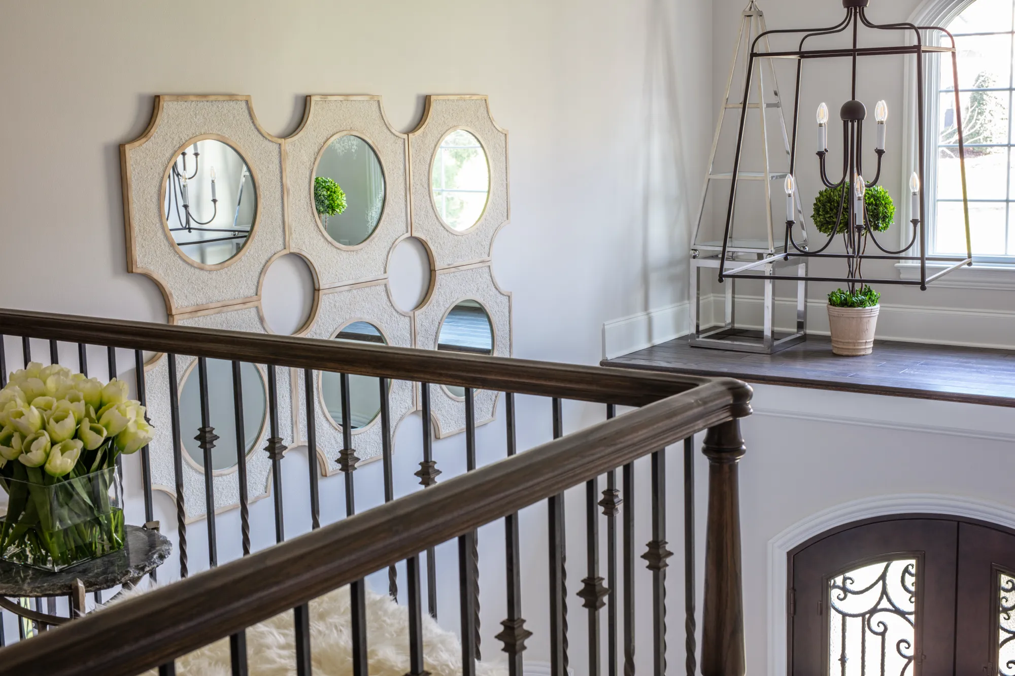 An elegant staircase landing features a dark wood railing, a multi-panel decorative mirror, and a modern black cage chandelier near an arched window.