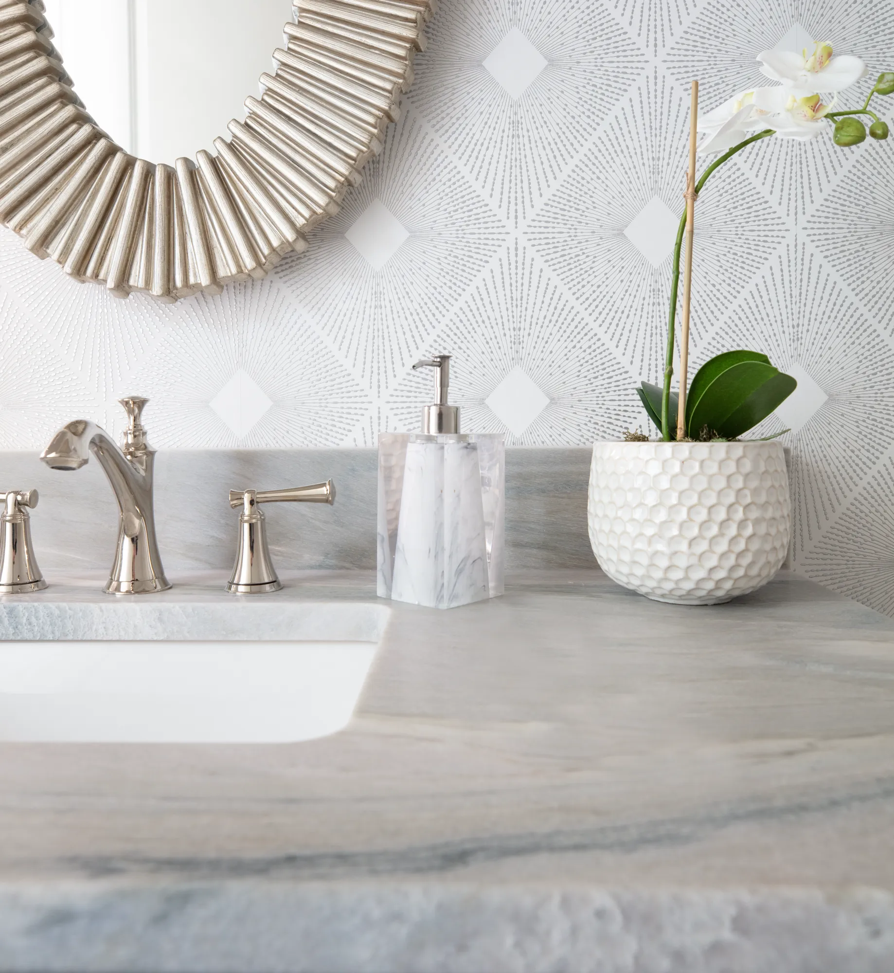 A close-up of a luxurious bathroom vanity features a marble countertop and a polished nickel faucet set. A marble-patterned soap dispenser and a white orchid in a textured pot sit against a gray starburst wallpaper.