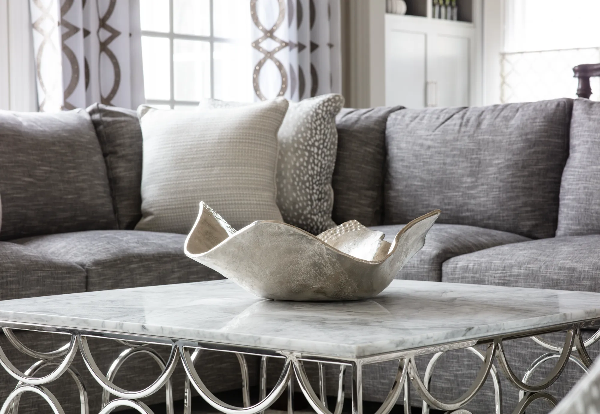 A close-up of a stylish gray sectional sofa and a marble coffee table with a chrome base. The table holds a reflective, silver centerpiece bowl.