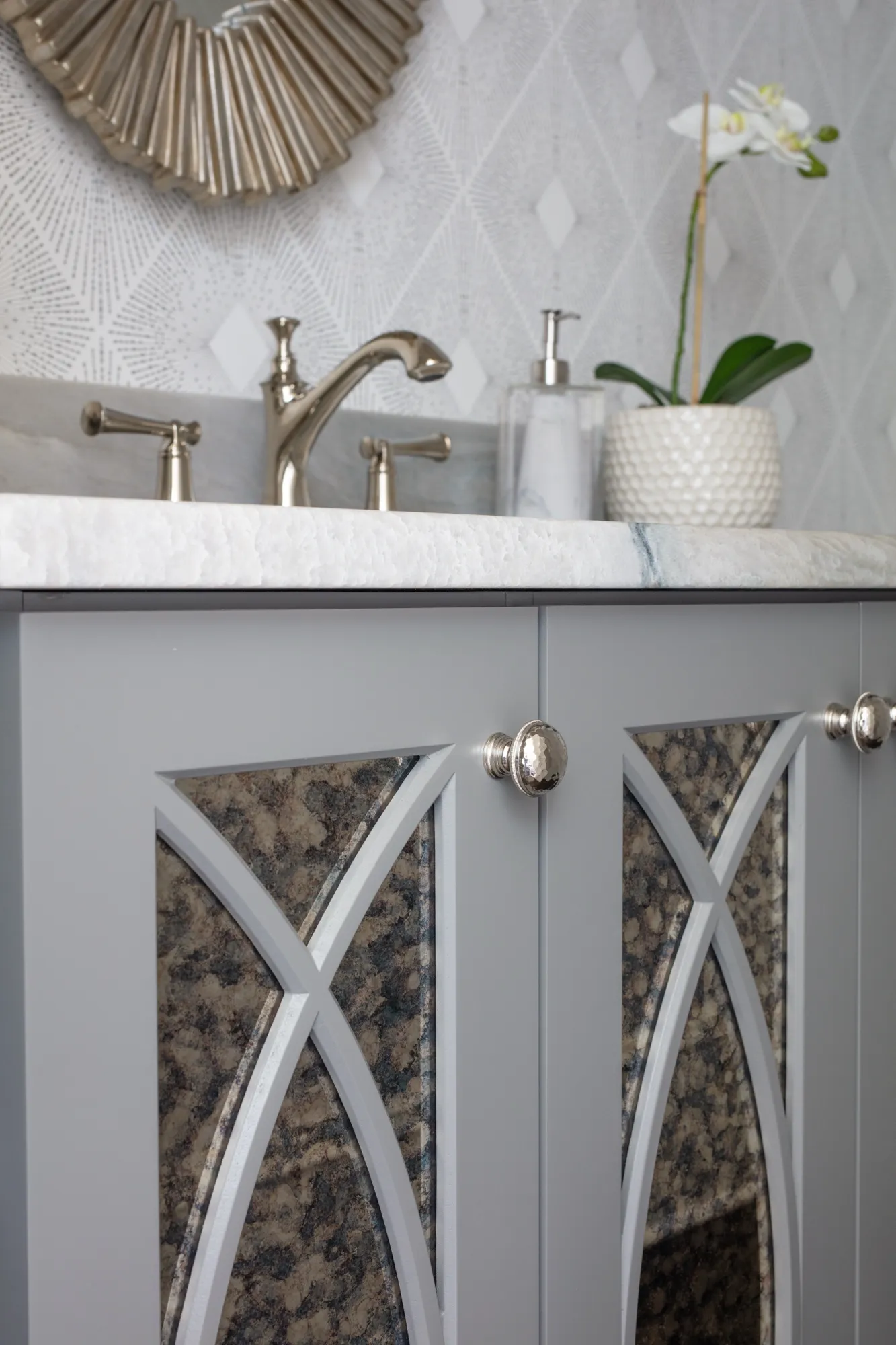 A modern gray vanity features a white marble top, silver faucet, and decorative cabinet doors over dark, mottled panels. A white orchid adds detail.